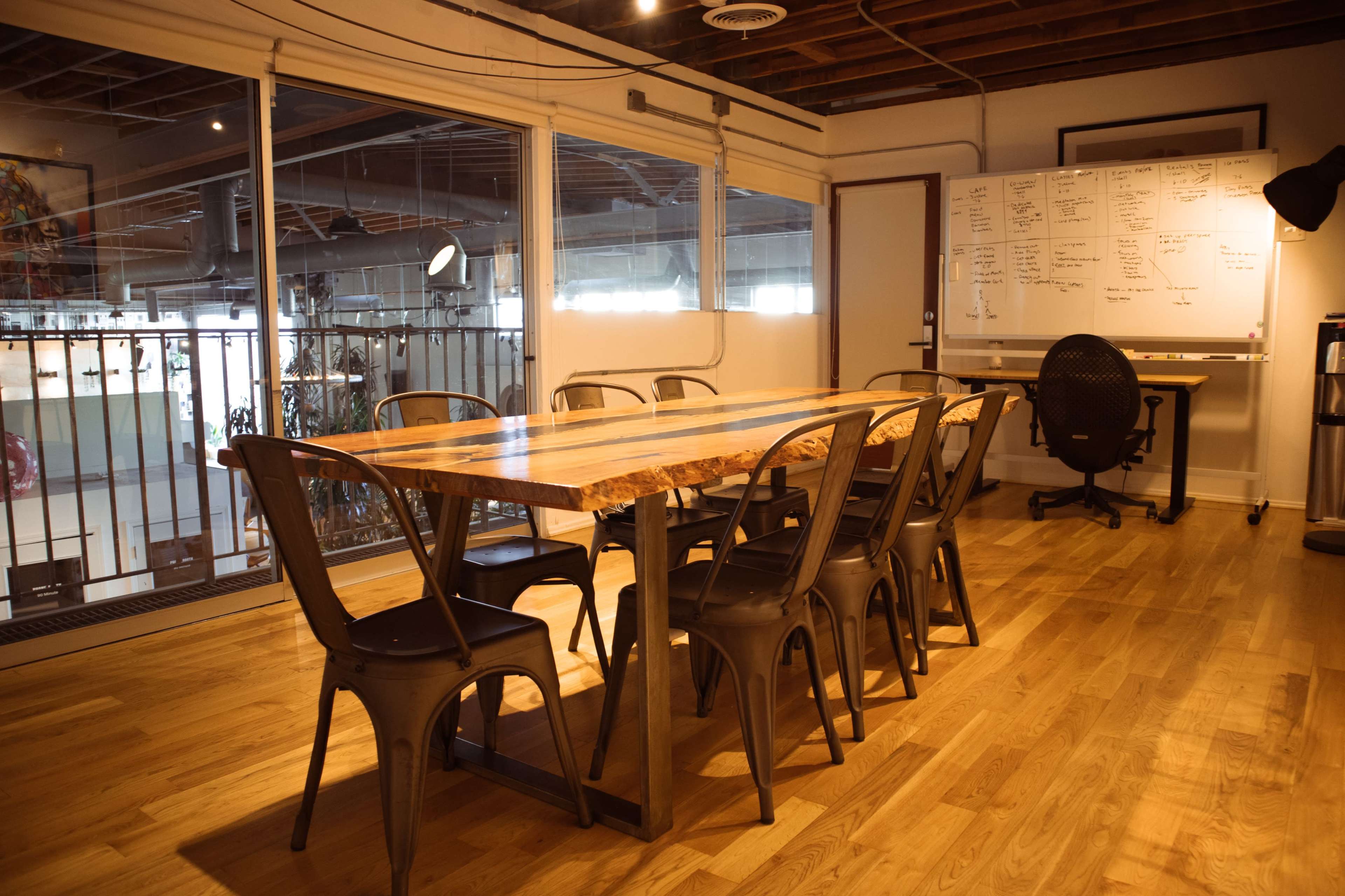 A large wooden table surrounded by metal chairs is positioned in a well-lit room with large windows and a whiteboard nearby.