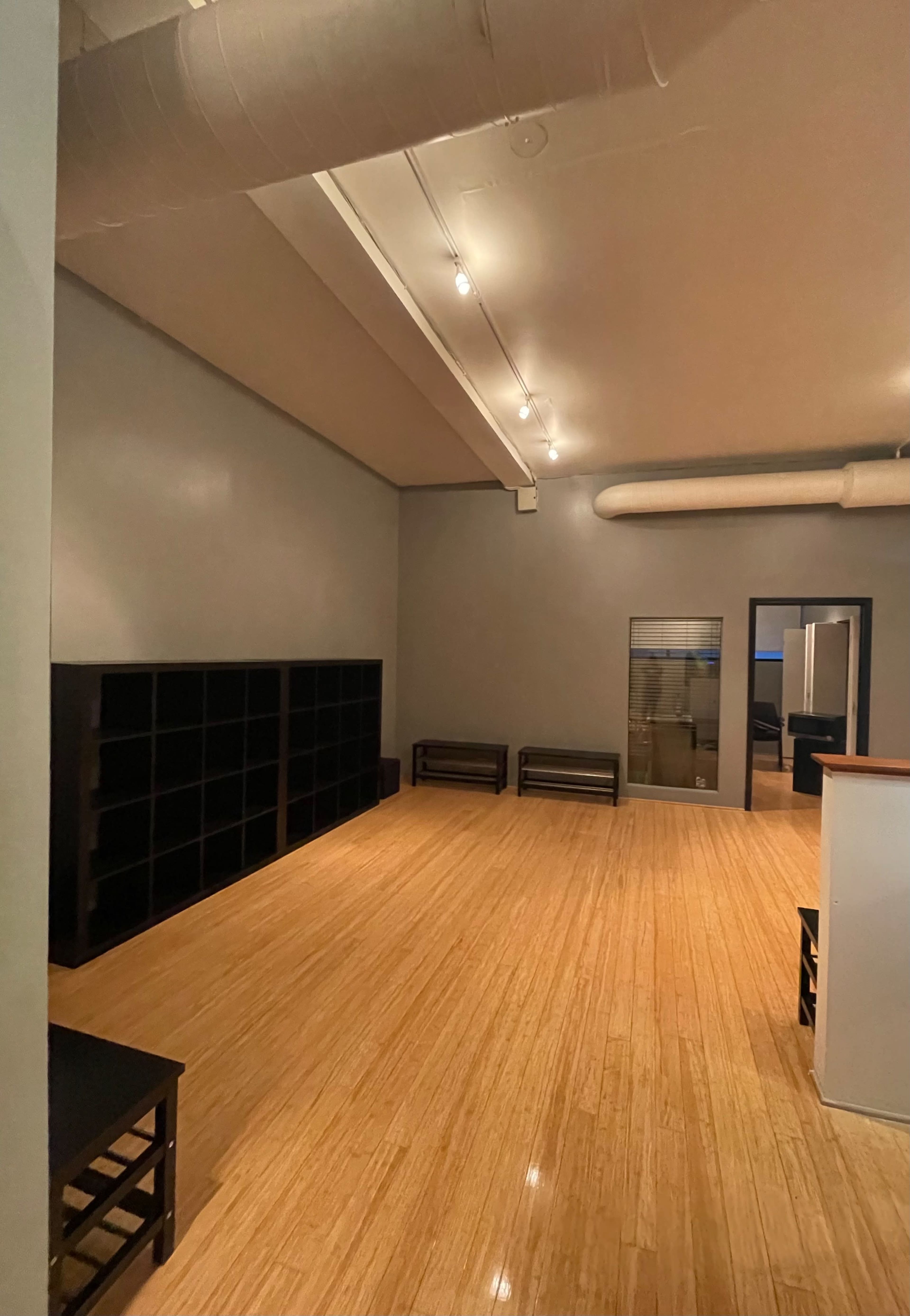 The image shows a spacious, empty room with bamboo flooring, gray walls, and shelving units along one side.