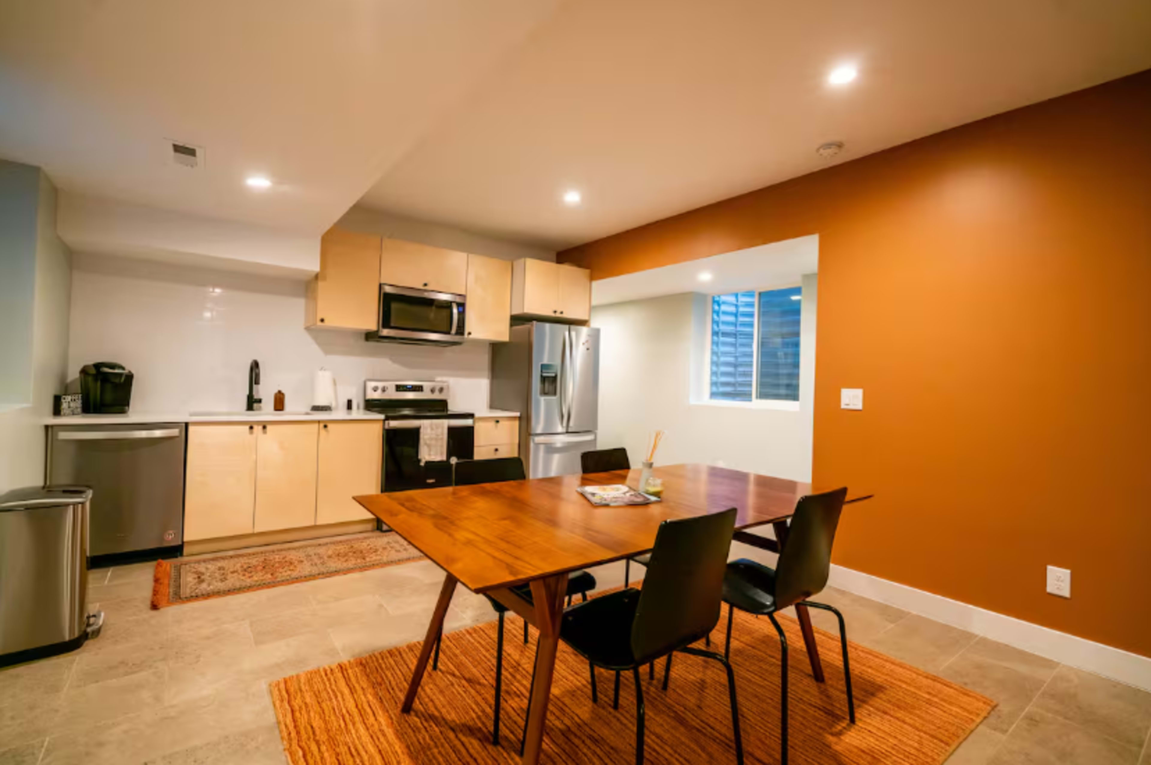 The image shows a modern kitchen and dining area with a wooden table, black chairs, and appliances, set against a brown accent wall.