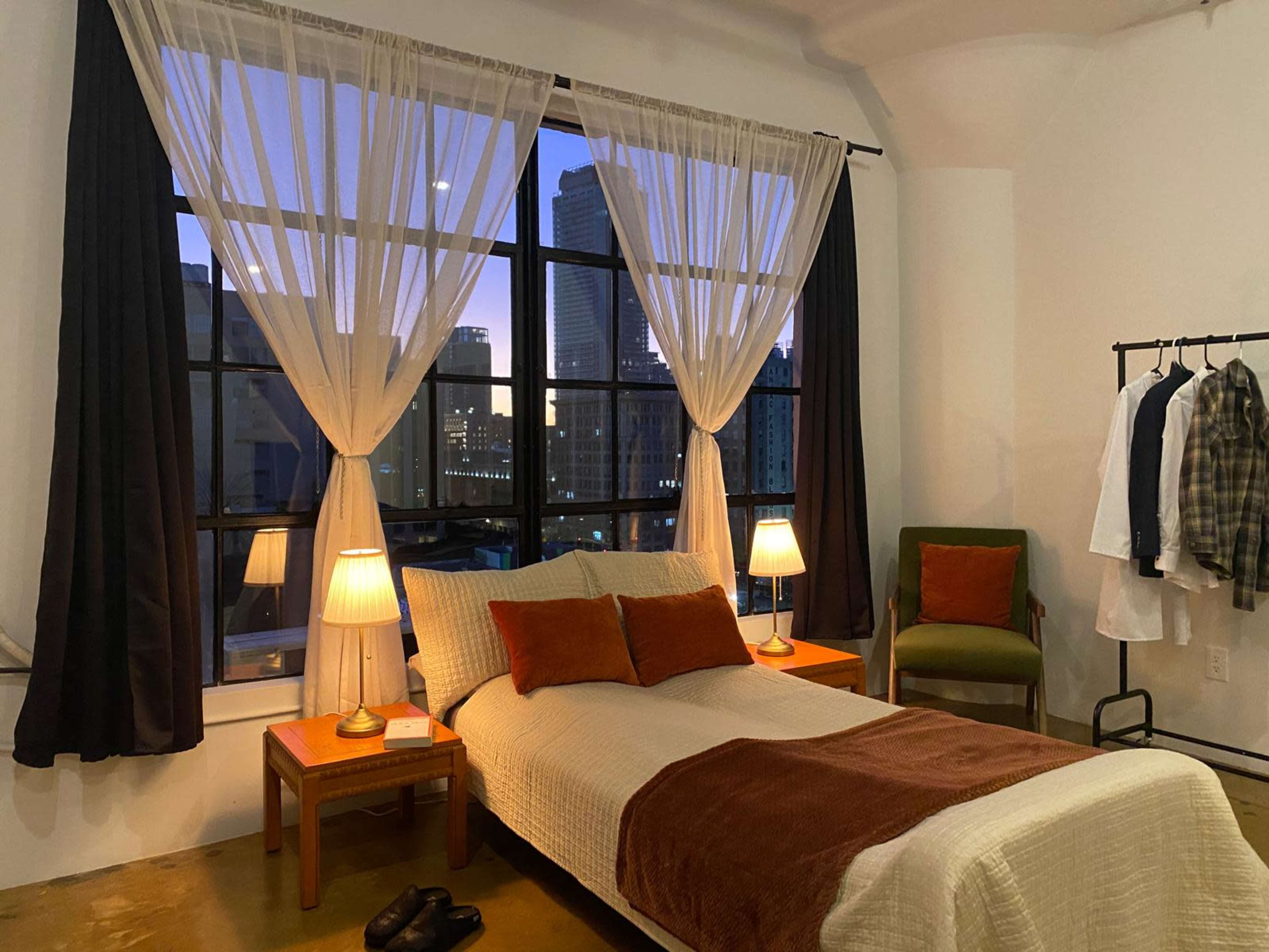 The image shows a styled bedroom with a bed, two lamps on side tables, large windows revealing a city skyline at dusk, and a clothing rack.