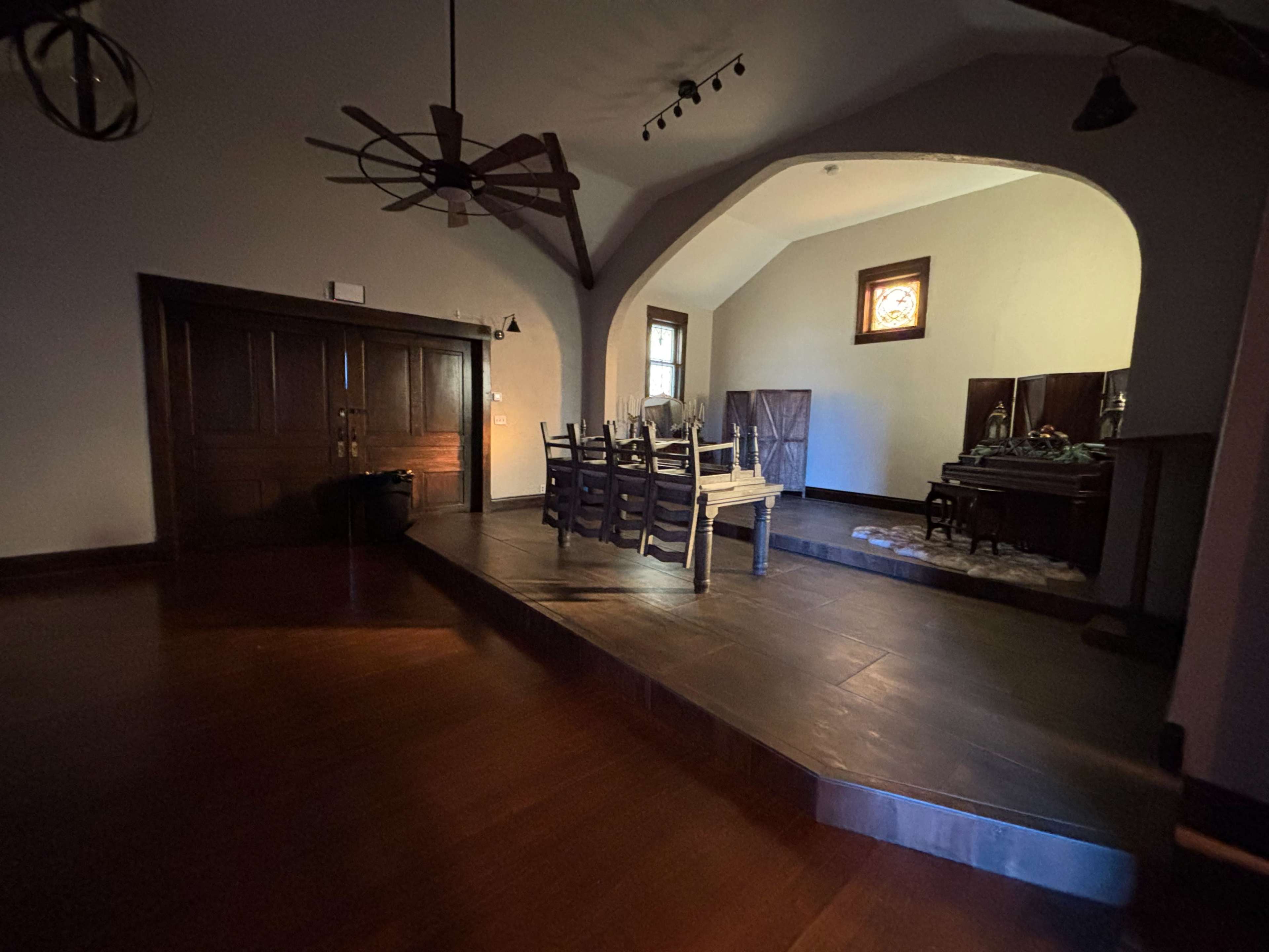 The image shows a partially lit dining area with a wooden table surrounded by chairs, located on a raised platform beneath an arched ceiling.