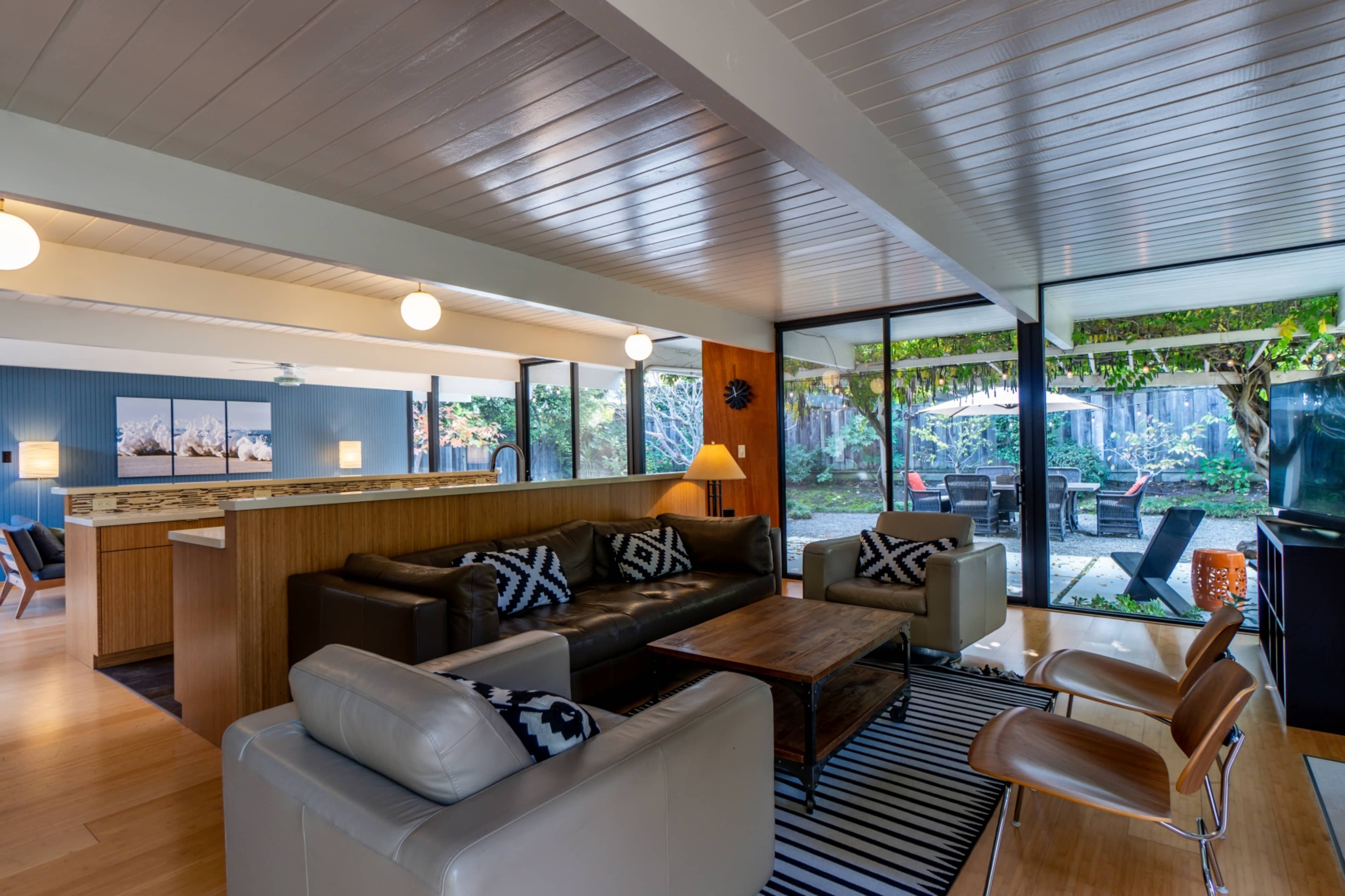 The image shows a modern, open-concept living room with seating arrangements, wooden accents, and large glass windows that overlook a backyard patio.