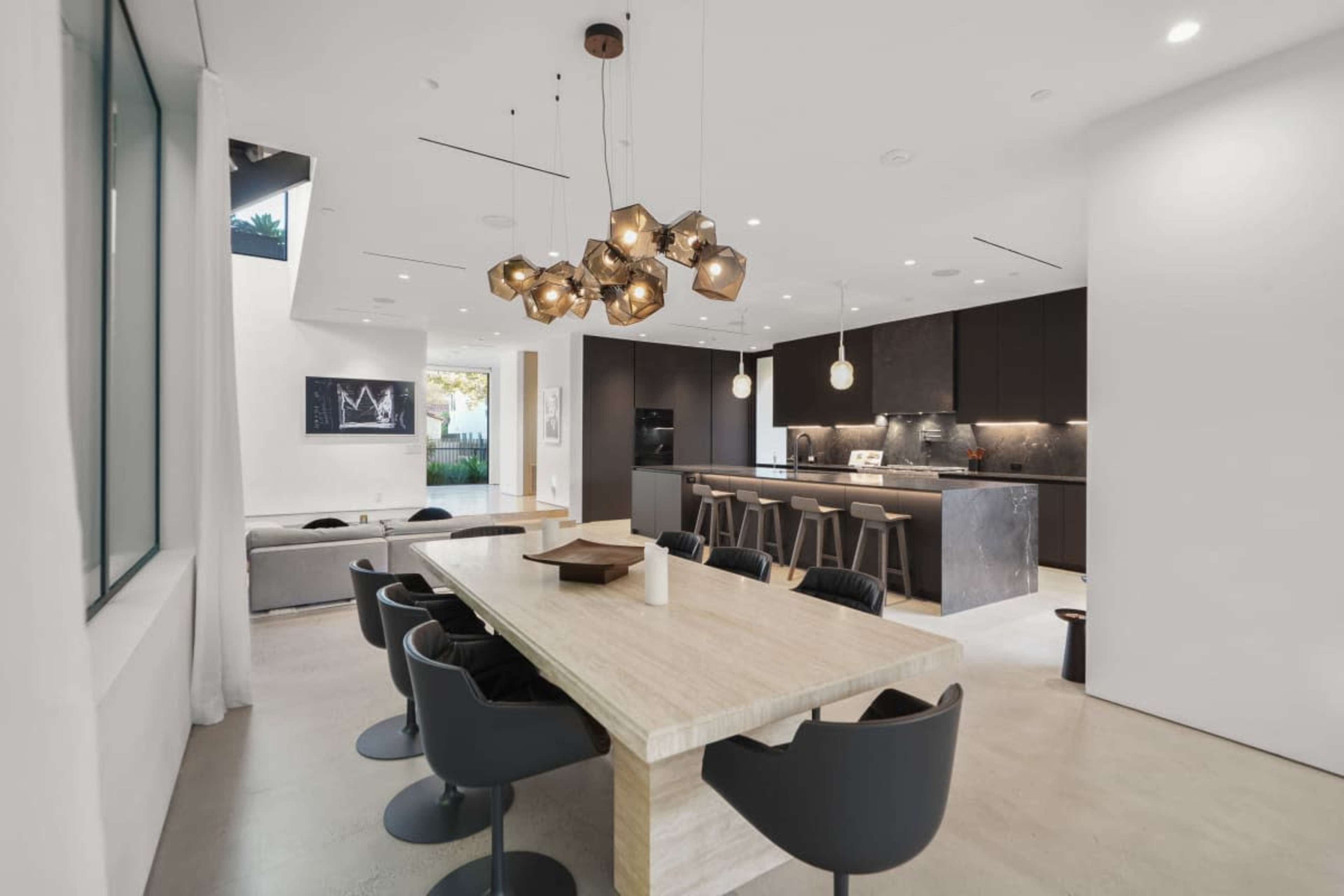The image shows a modern dining area featuring a large stone table surrounded by black chairs, with an open kitchen visible in the background.