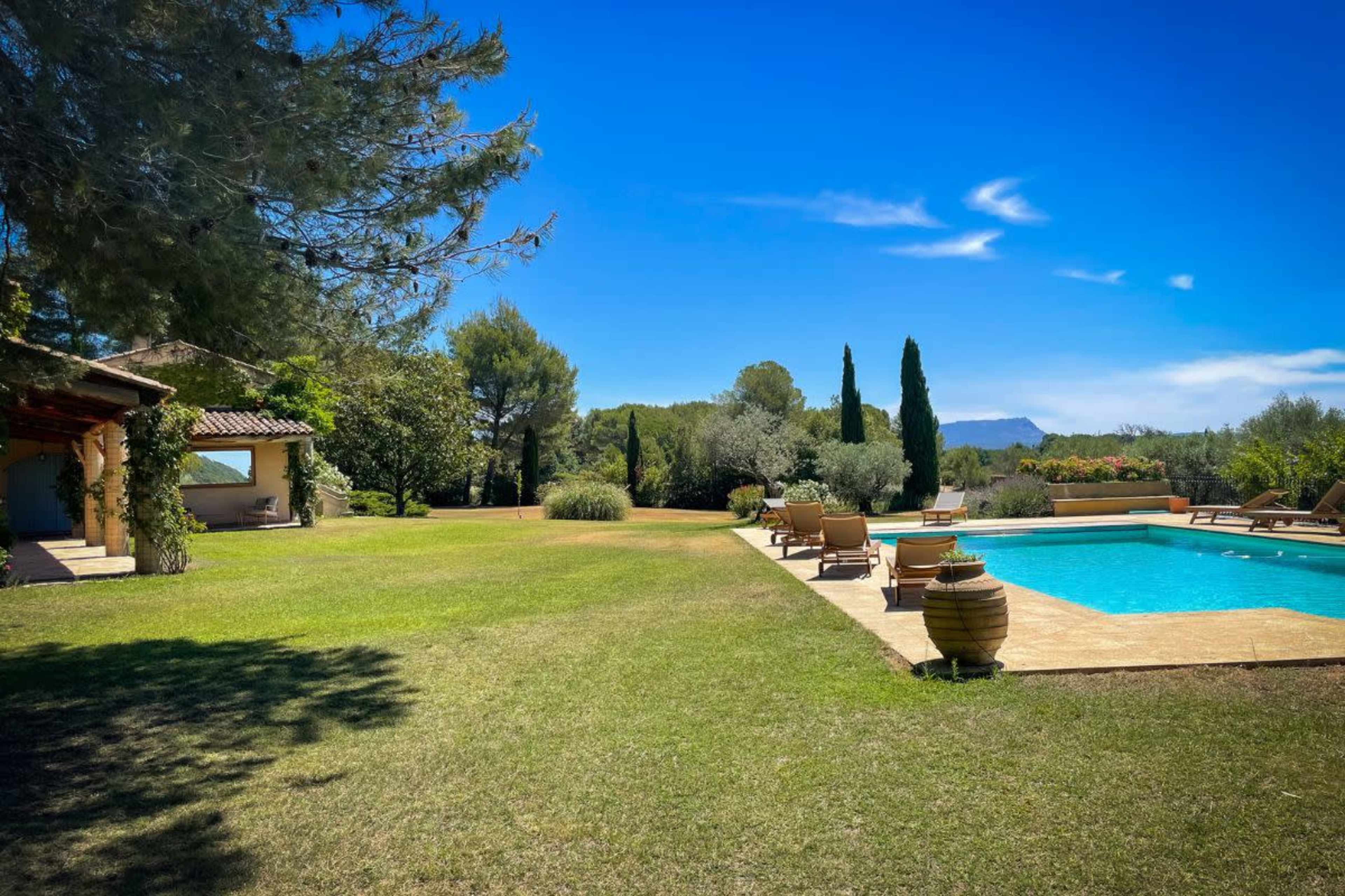 A landscaped backyard featuring a swimming pool surrounded by lounge chairs, with trees and a distant mountain in the background under a clear blue sky.