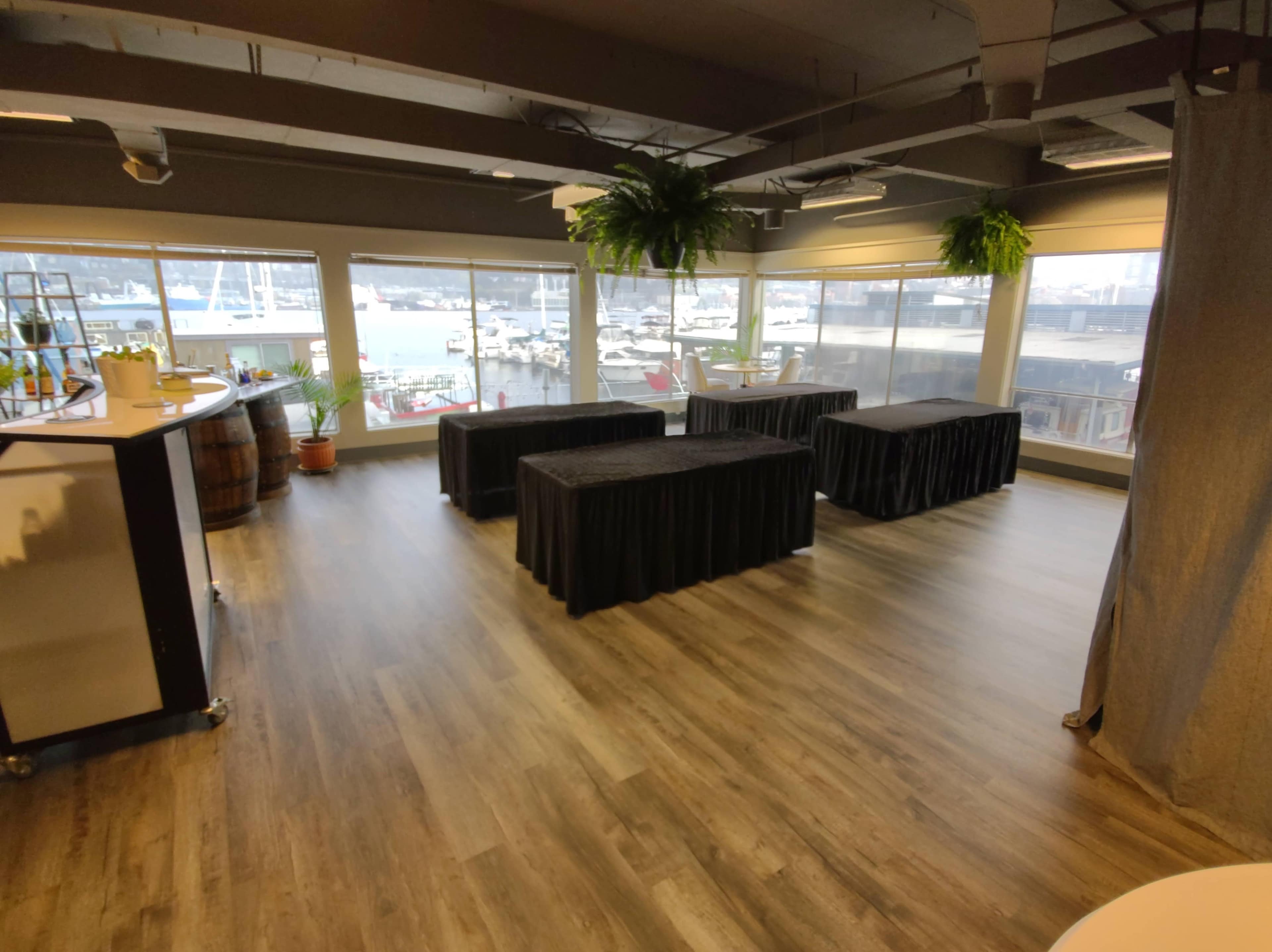 The interior space features several tables covered with black tablecloths, large windows showcasing a view of boats outside, and a bar area with decorative plants.