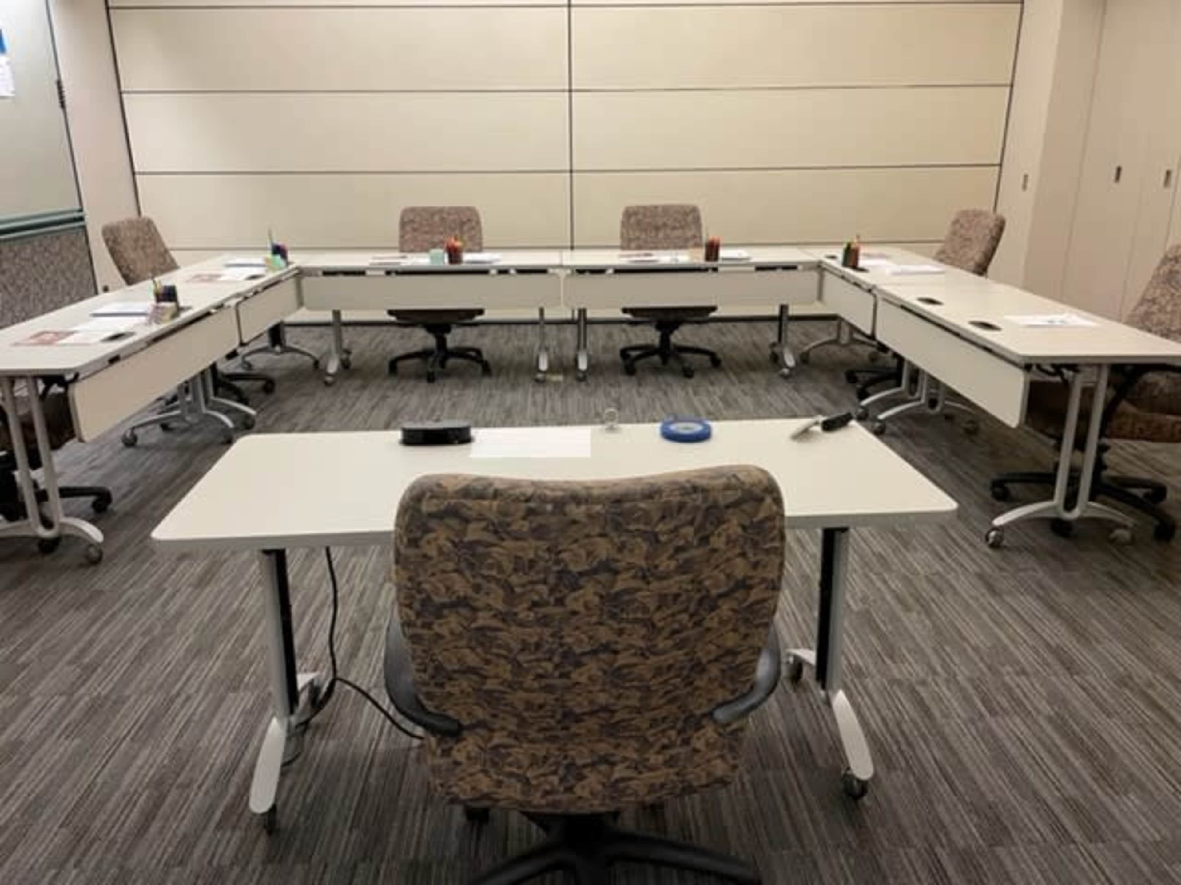 A conference room is set up with multiple tables arranged in a U-shape, with chairs and supplies visible at each station.