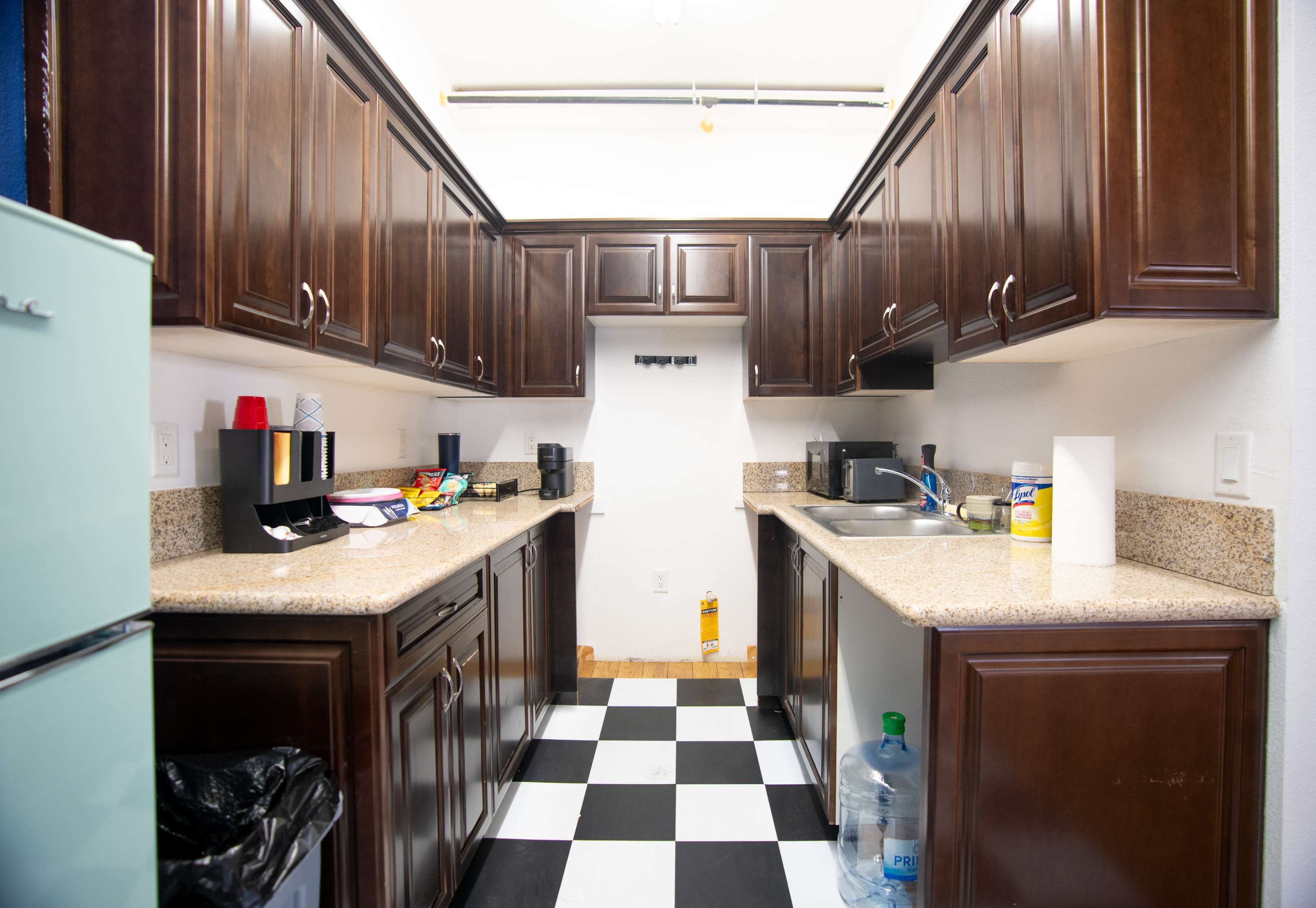 The image shows a narrow kitchen with dark wooden cabinets, light granite countertops, and a black and white checkered floor.