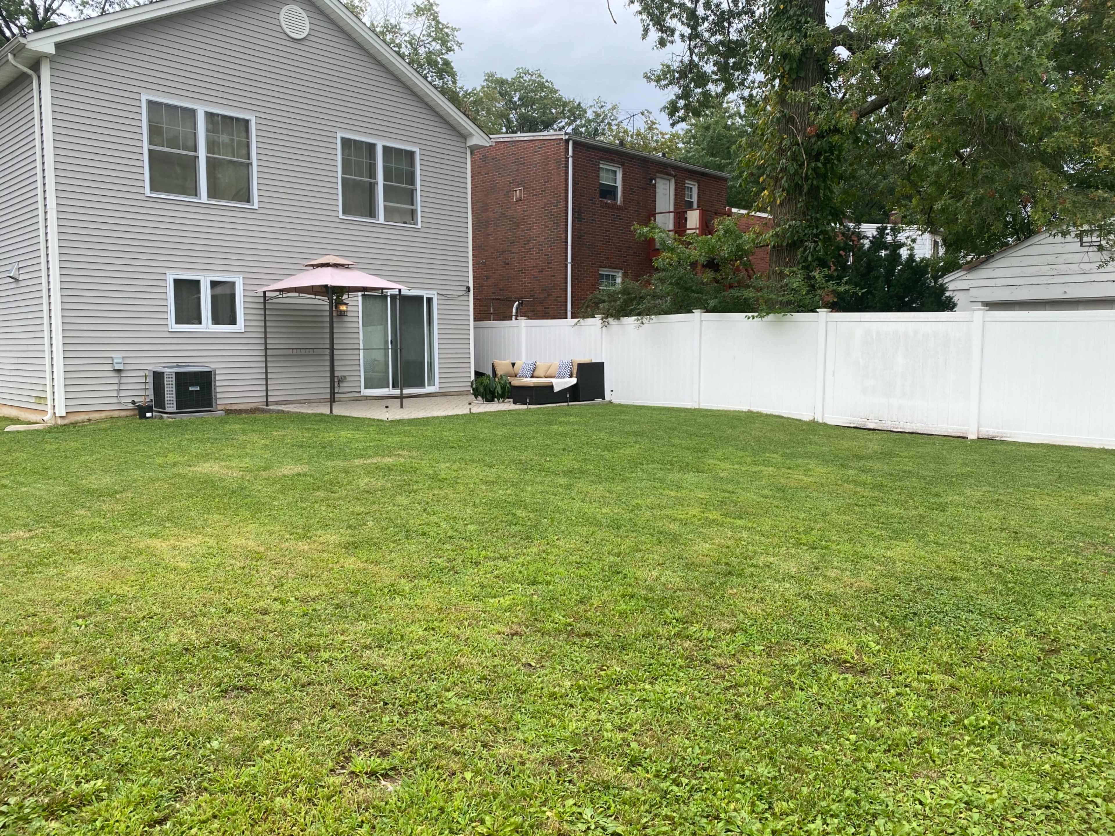 A neatly maintained backyard features a lawn, a gray house with large windows, a patio area with seating, and a white privacy fence.