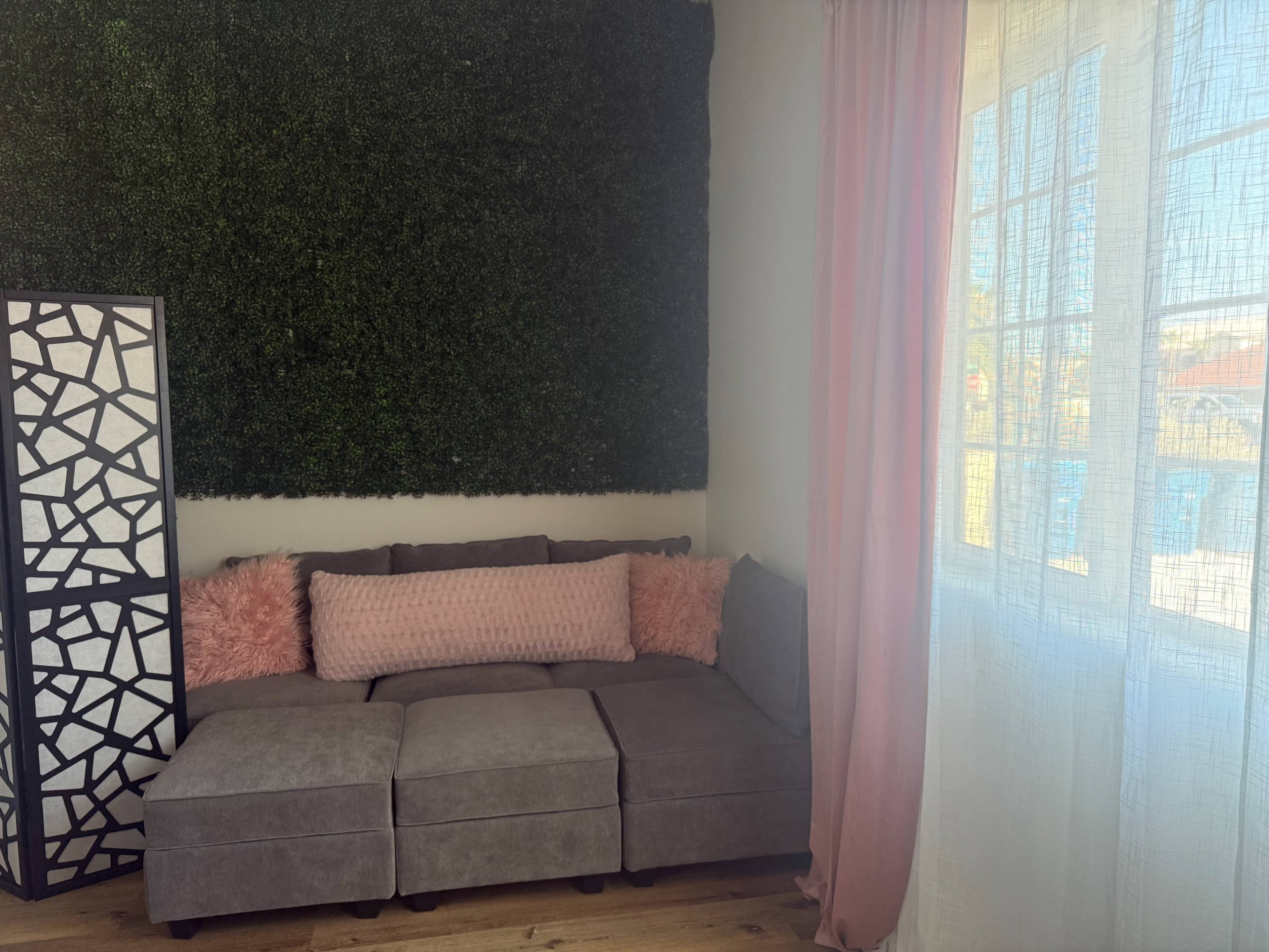 The image shows a cozy corner of a room featuring a gray sectional sofa with pink decorative pillows, a tall black and white screen, and a green artificial grass wall backdrop.