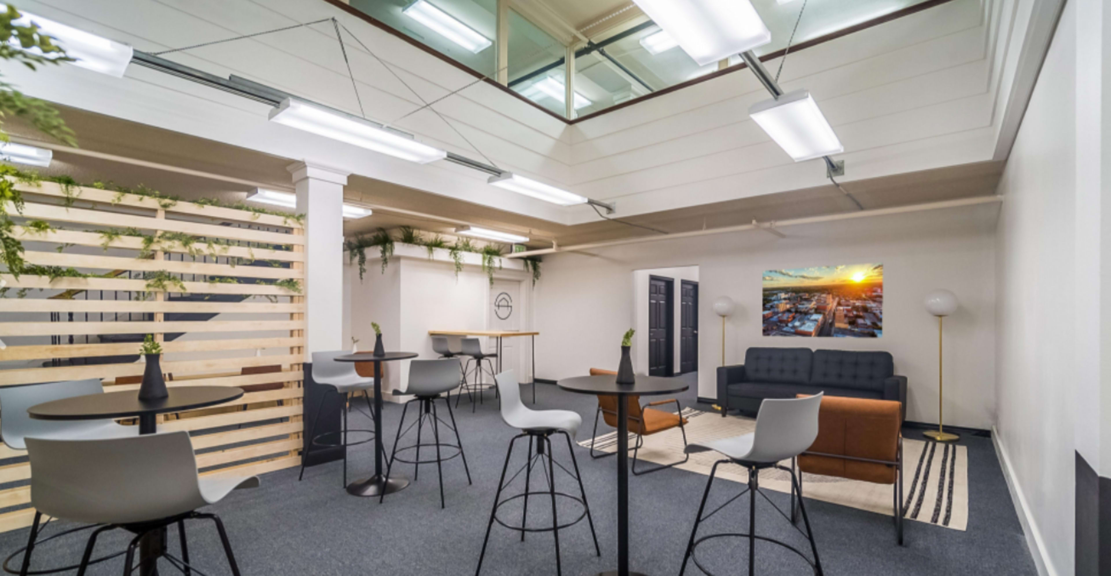 A modern office lounge area with several high tables and chairs, a couch, plants, and a large window providing natural light.