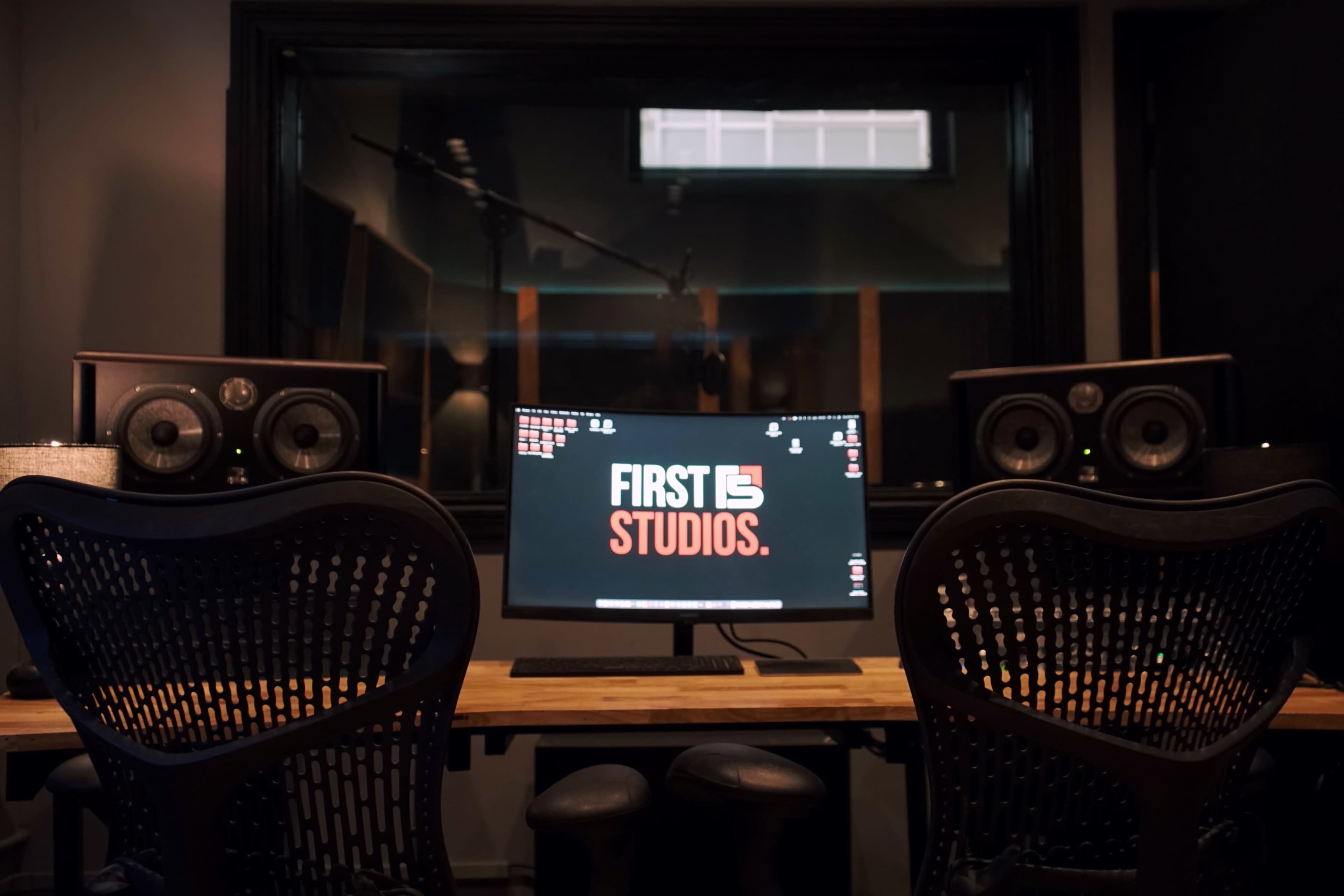 The image shows a recording studio with two chairs facing a computer displaying the text "FIRST 15 STUDIOS" and audio monitors on either side.