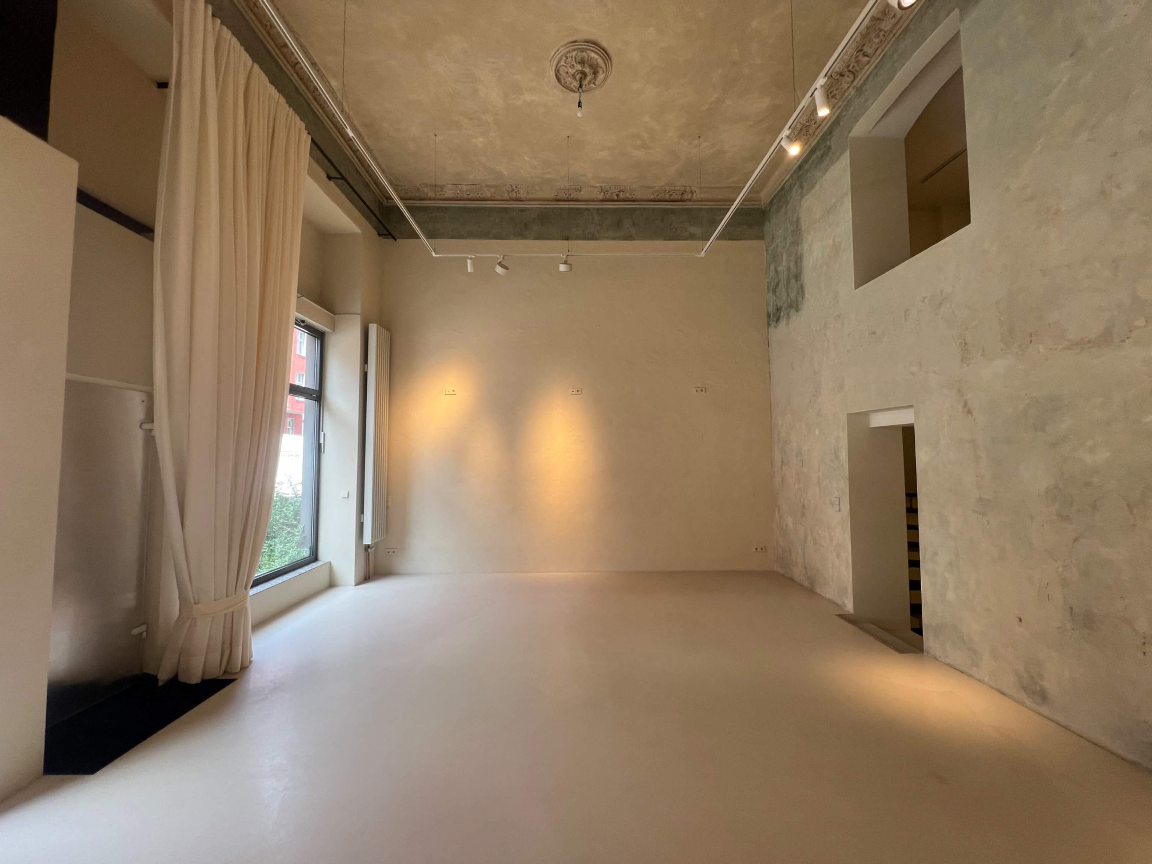 The image shows a spacious, empty room with high ceilings, a large window, and light-colored walls.