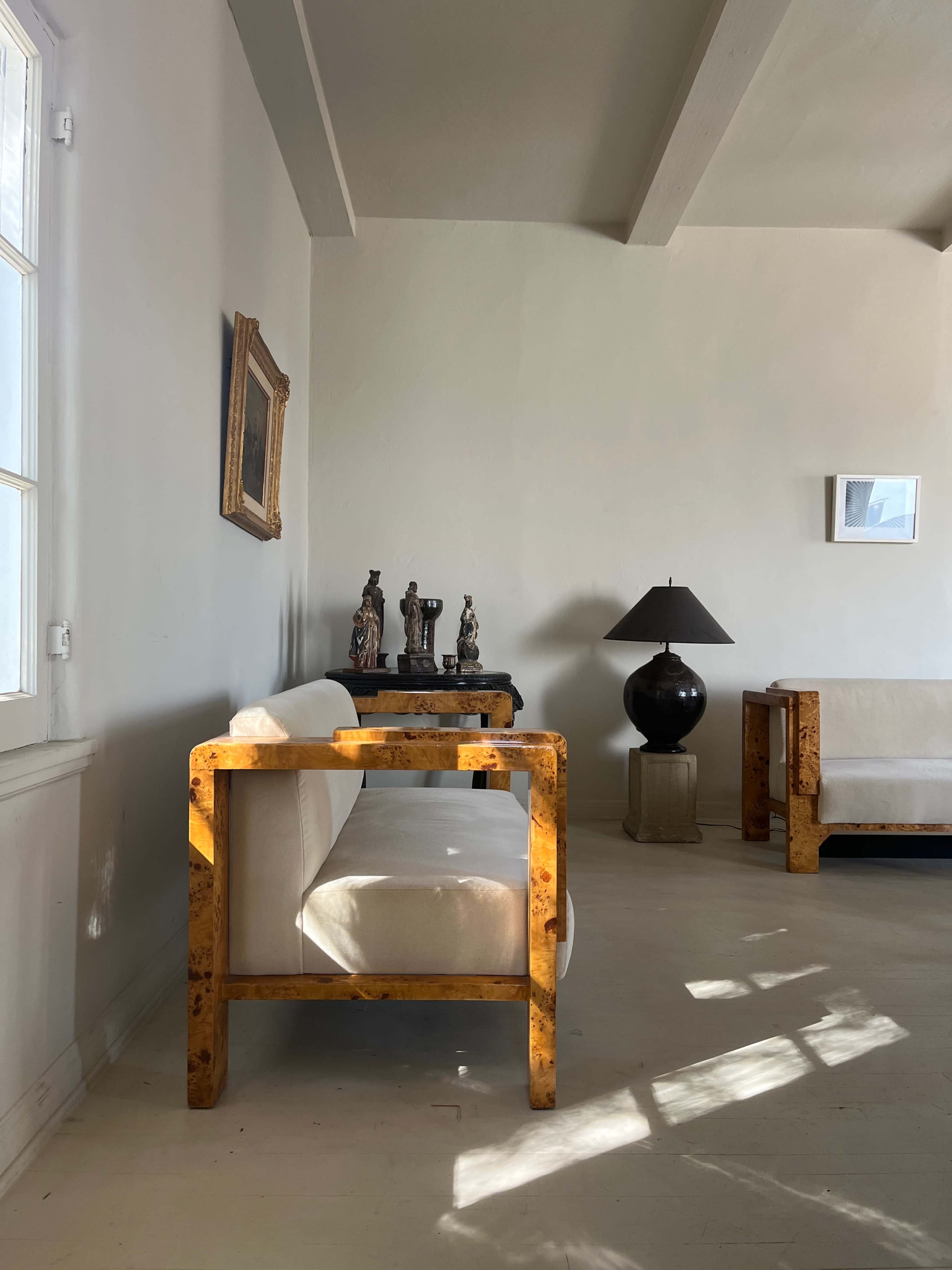 A minimalist living space features a light-colored sofa with wooden armrests, a black lamp, and several sculptures on a table against a white wall.