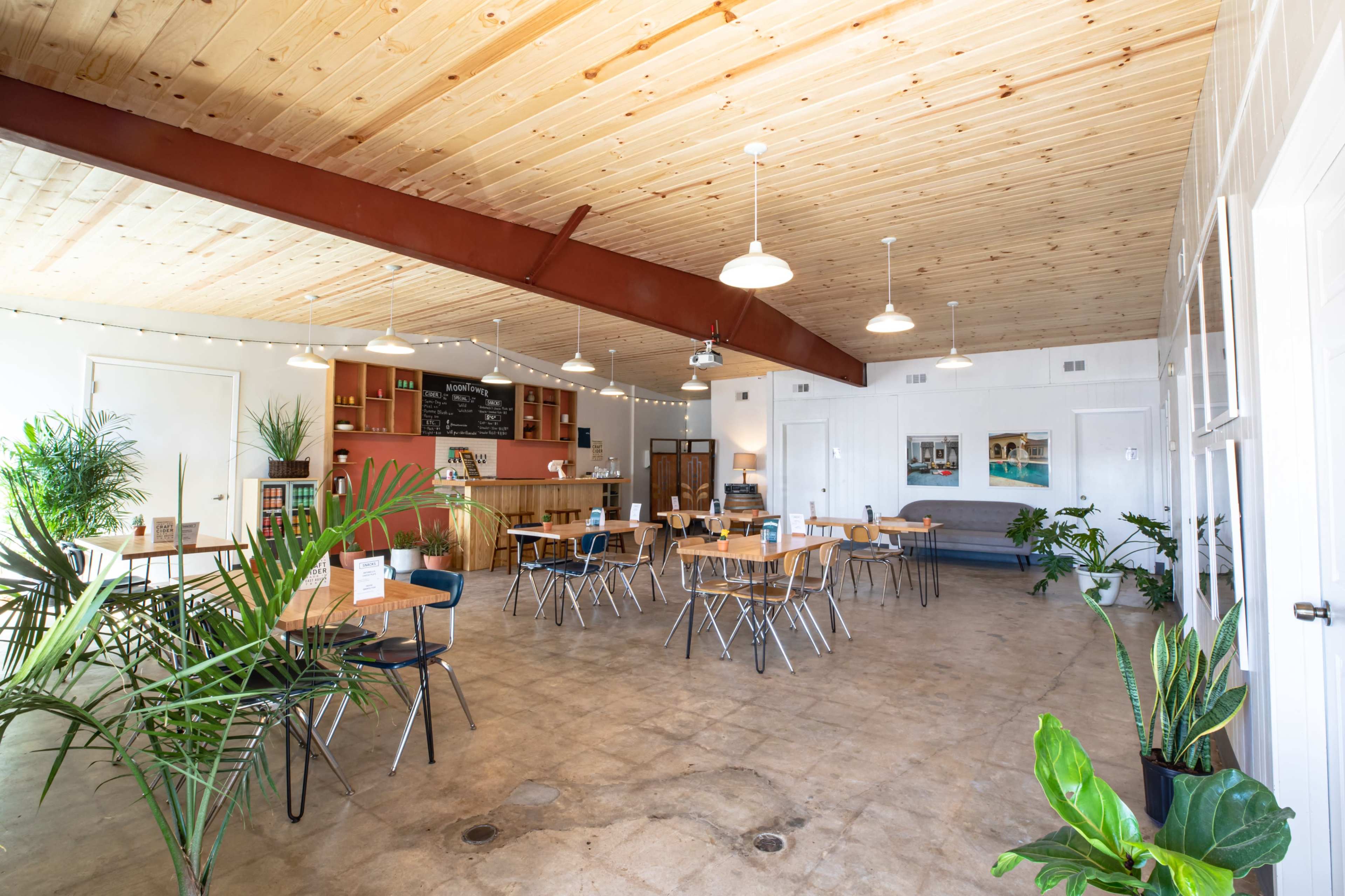 A spacious dining area with several wooden tables and chairs arranged around potted plants, feature lighting, and a counter in the background.