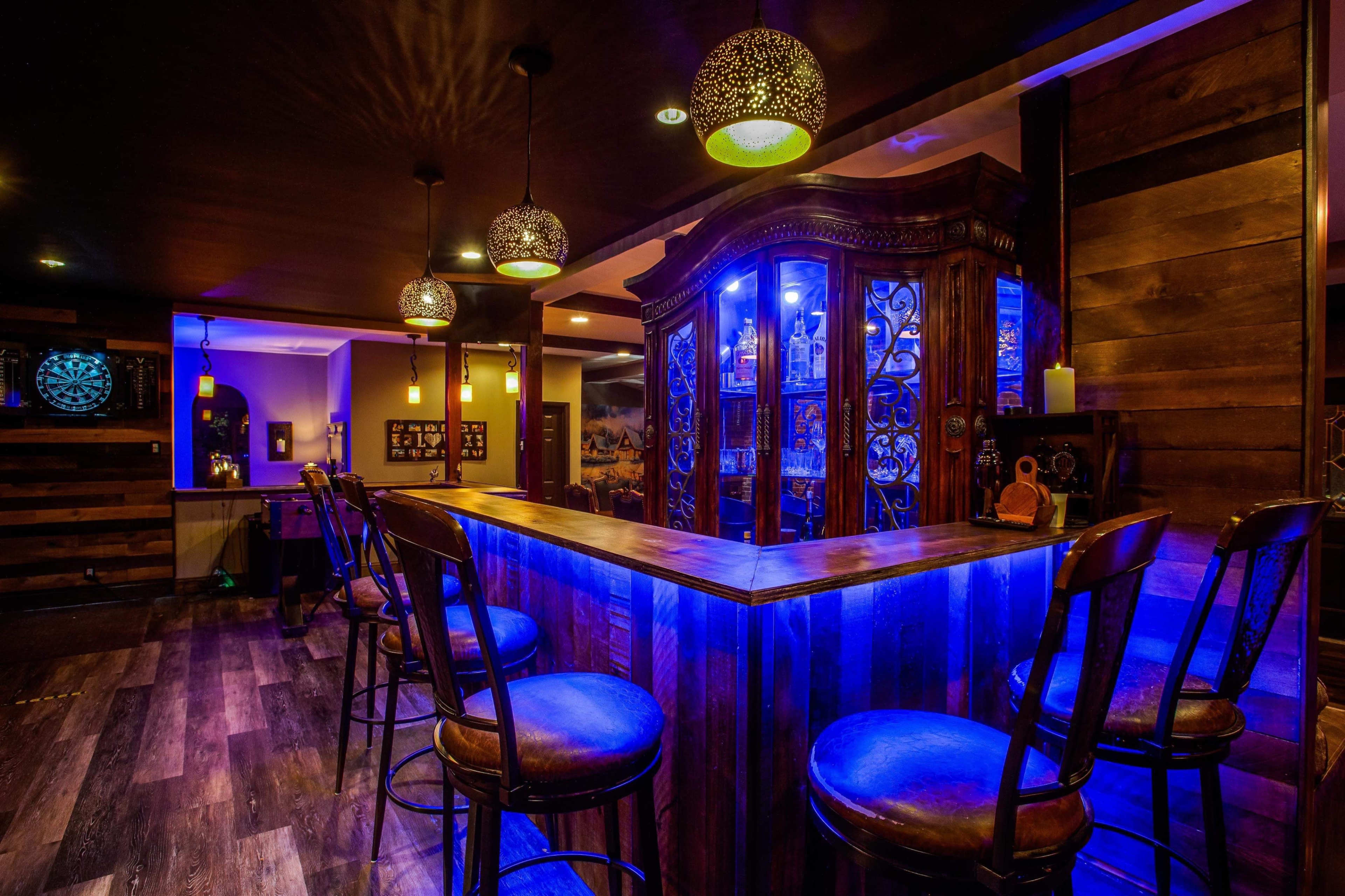 A well-lit bar area featuring a wooden counter and high chairs, illuminated by decorative pendant lights.