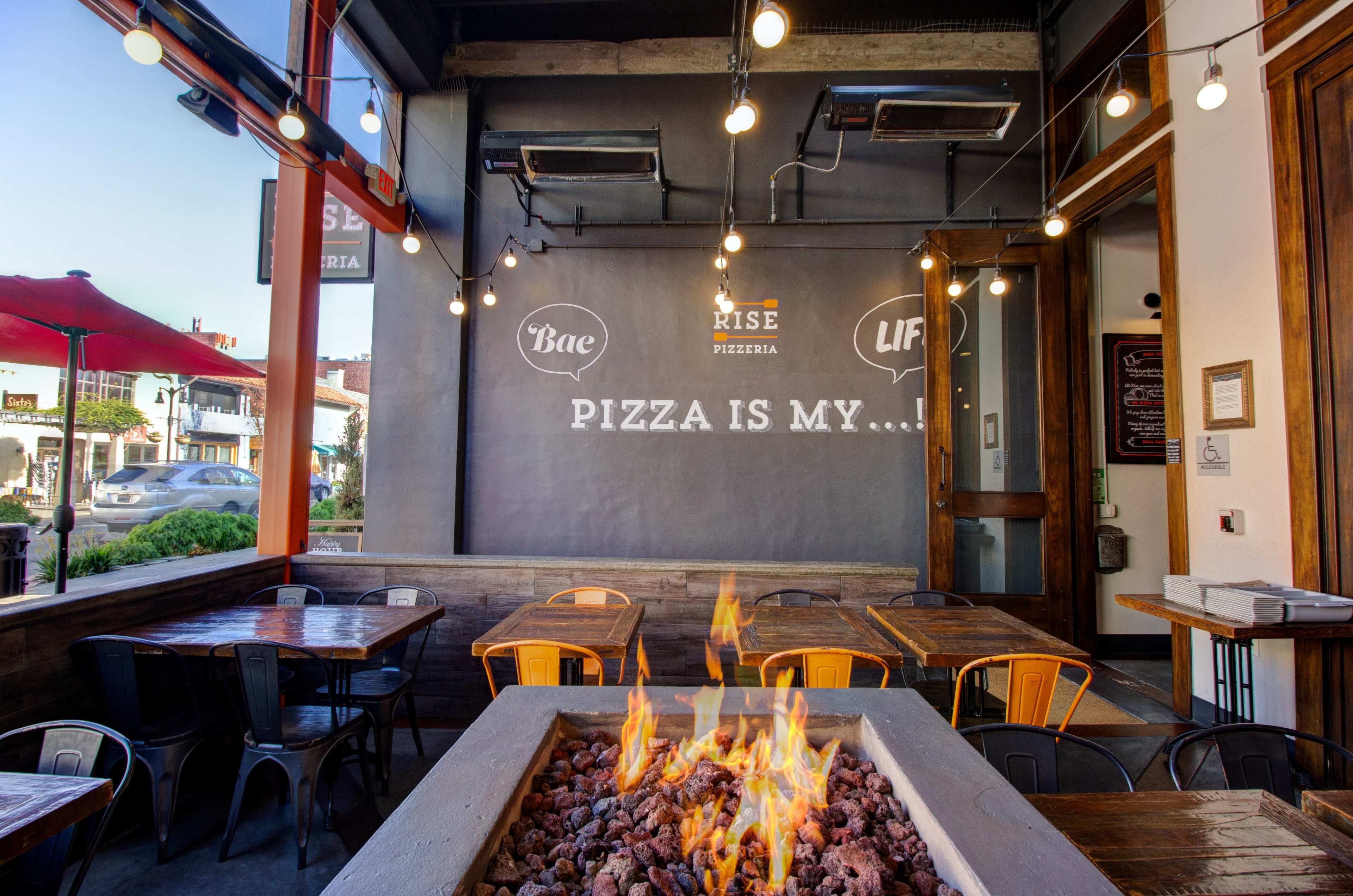 The interior of a pizzeria features a stone fire pit, wooden tables, and a wall with the words "PIZZA IS MY..." illuminated above.