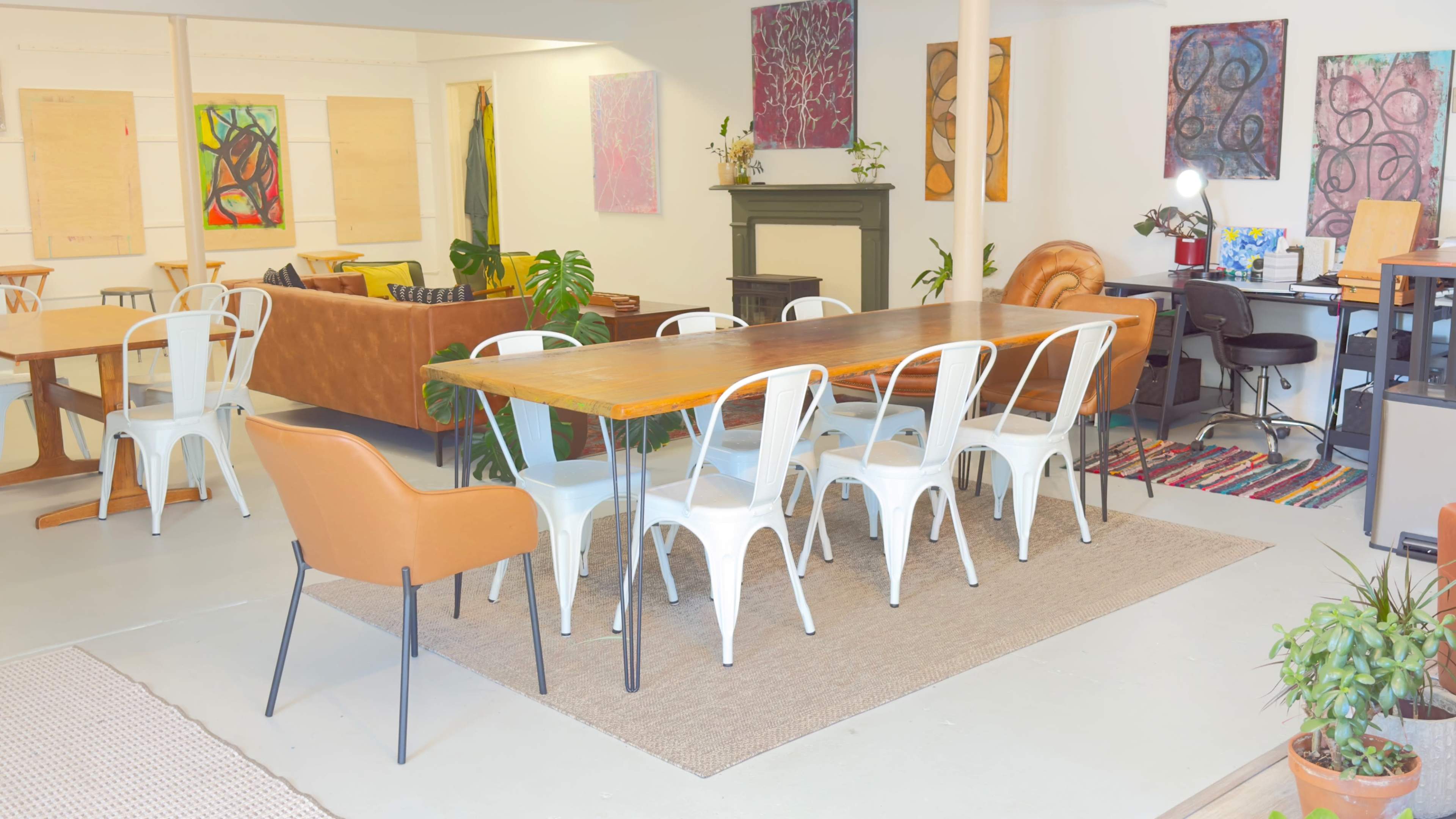 The image shows a spacious room featuring a long wooden dining table surrounded by white chairs, a large brown couch, decorative art pieces on the walls, and various plants.