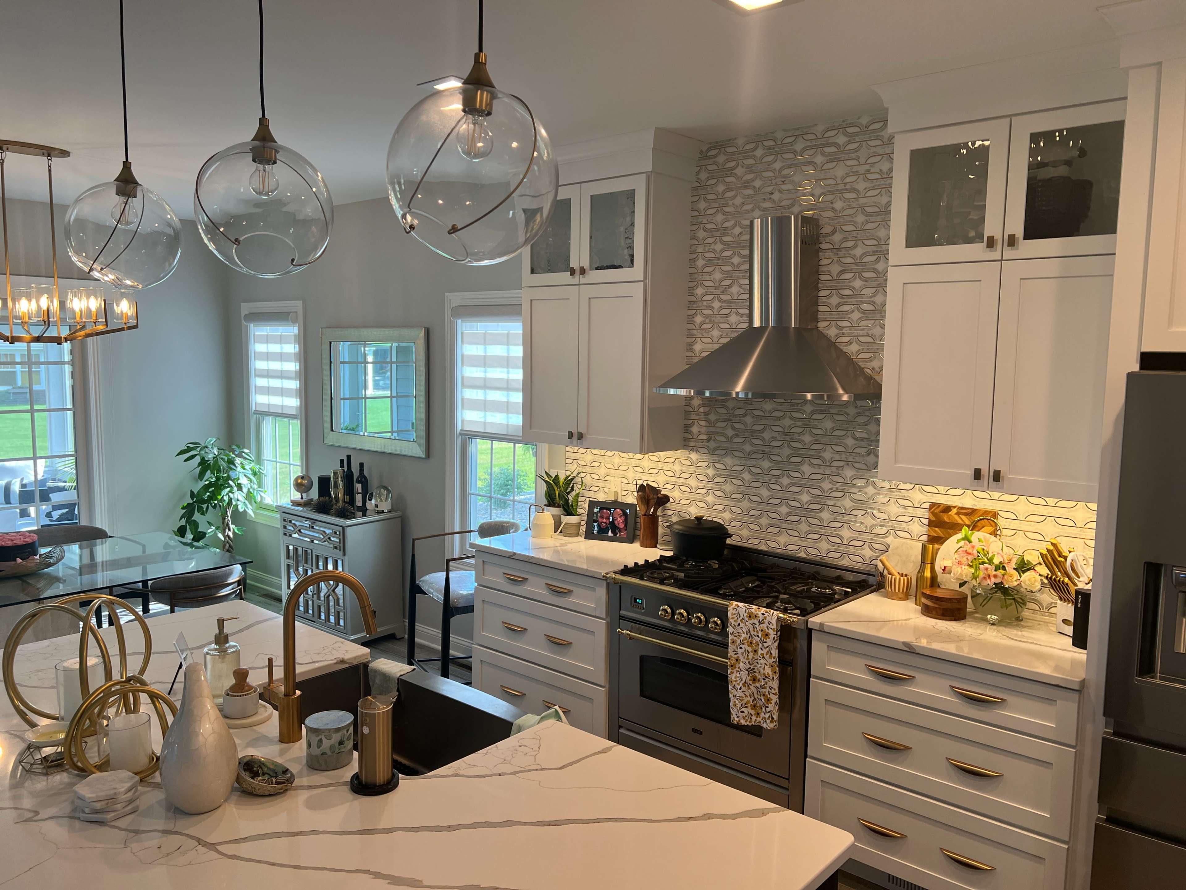 The image shows a modern kitchen featuring white cabinetry, a marble countertop, a gas stove with a hood, and large pendant lights hanging from the ceiling.