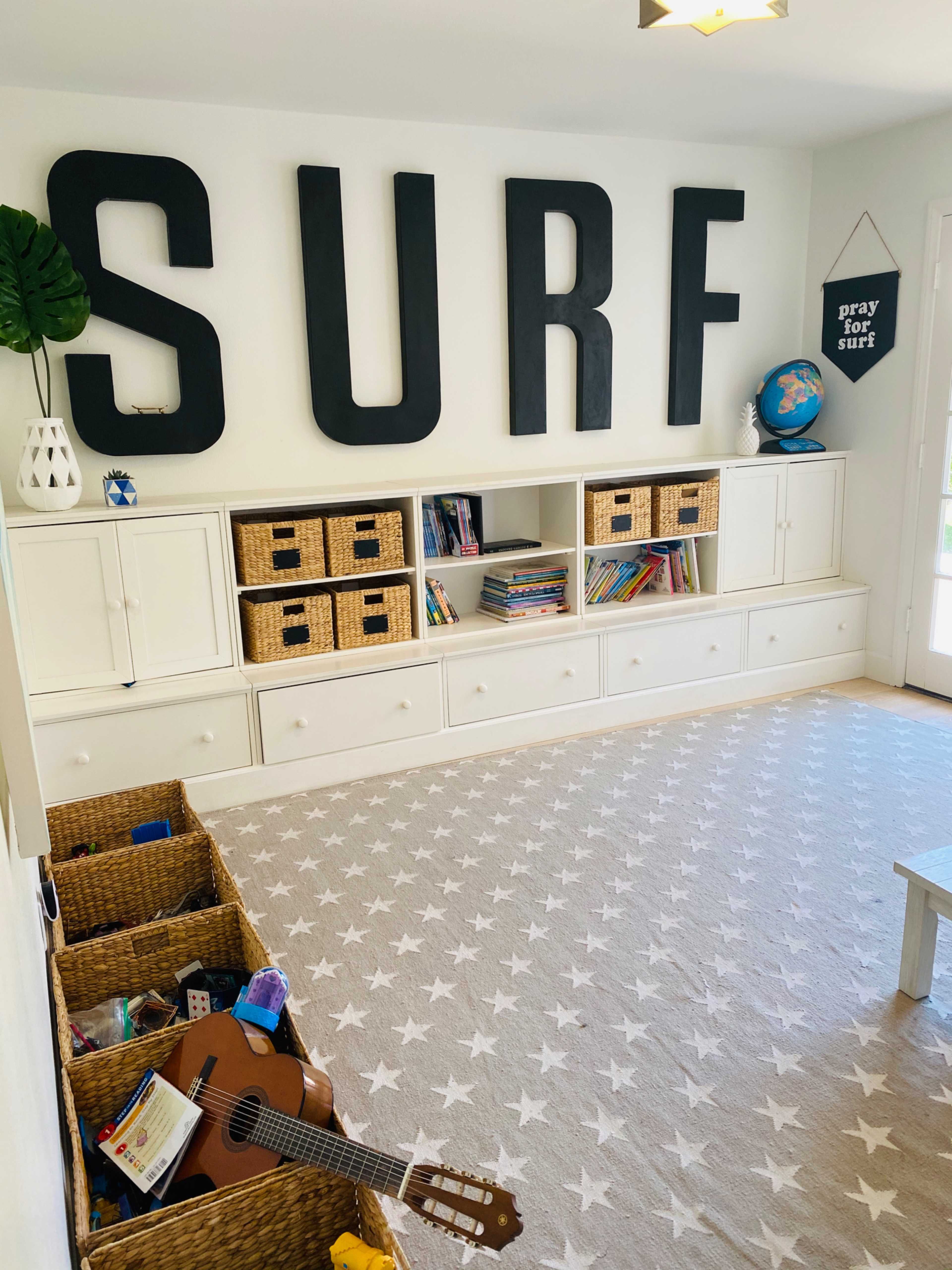 A playroom features large black letters spelling "SURF" on the wall, white storage units with woven baskets, shelves filled with books and toys, and a star-patterned rug.