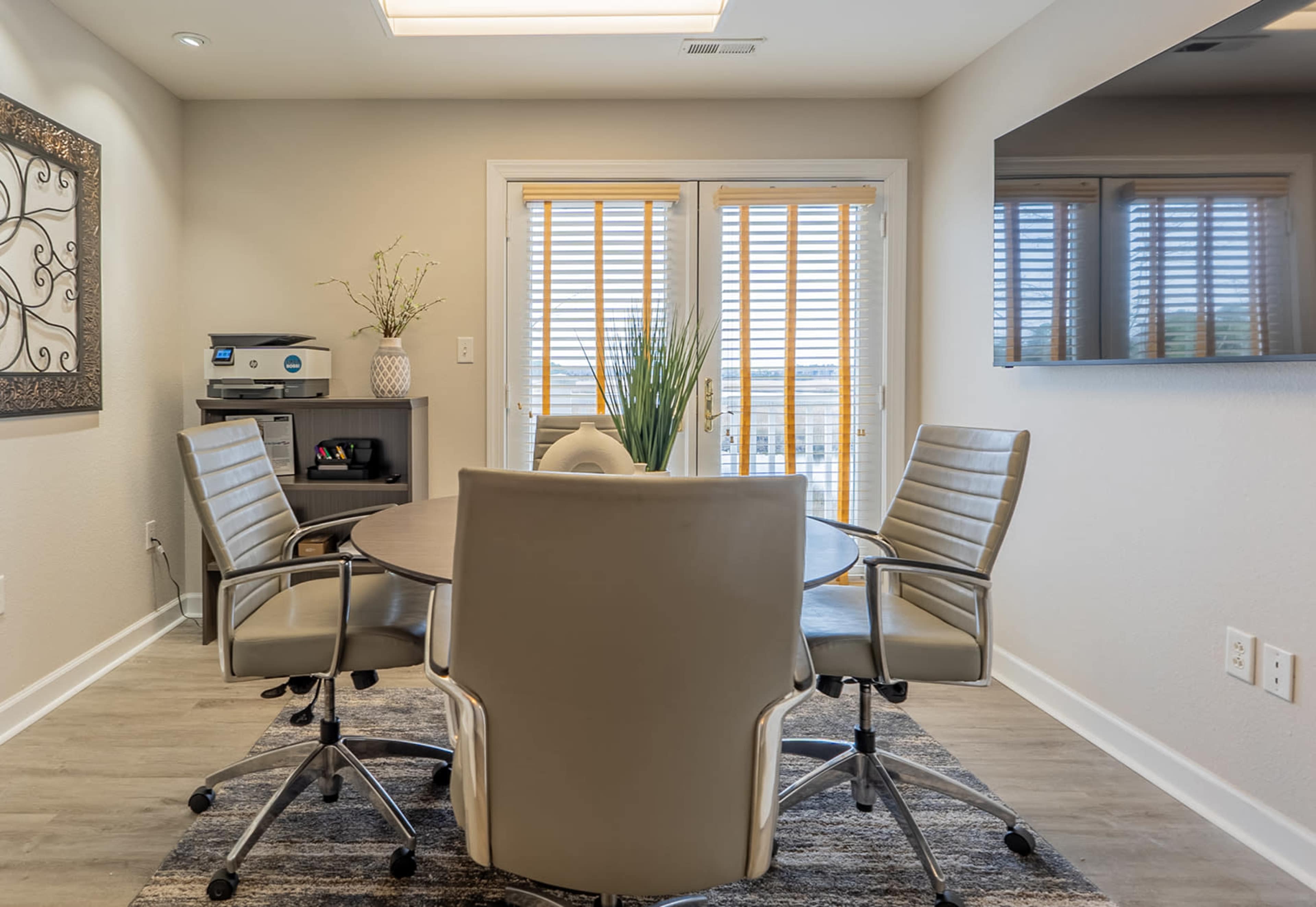 A modern meeting room features a round table surrounded by four ergonomic chairs, with a decorative wall, a storage unit, and large windows with blinds.