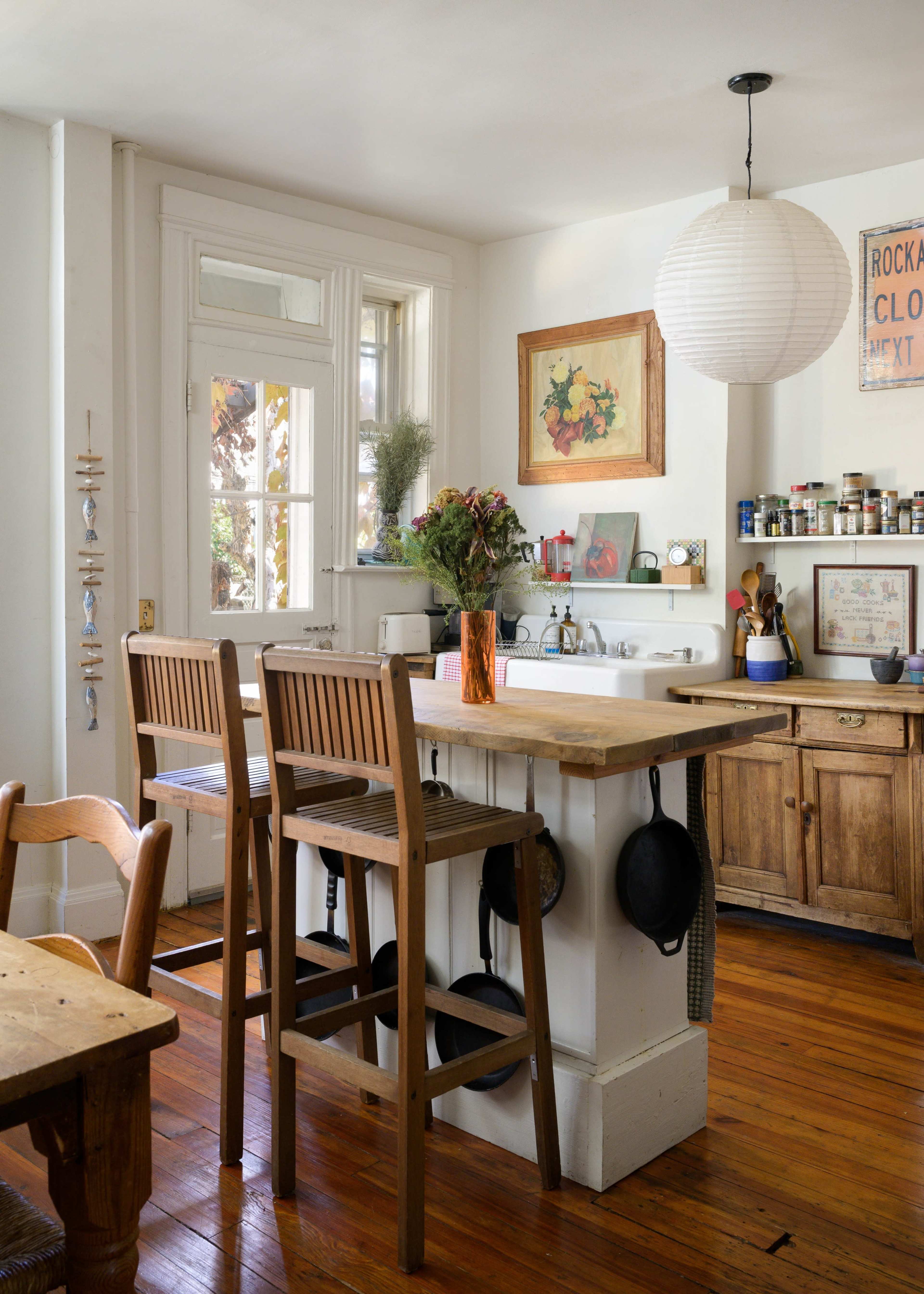 The image shows a bright kitchen with wooden bar stools around a small island, featuring plants, decorative art, and organized shelves with various spices and utensils.