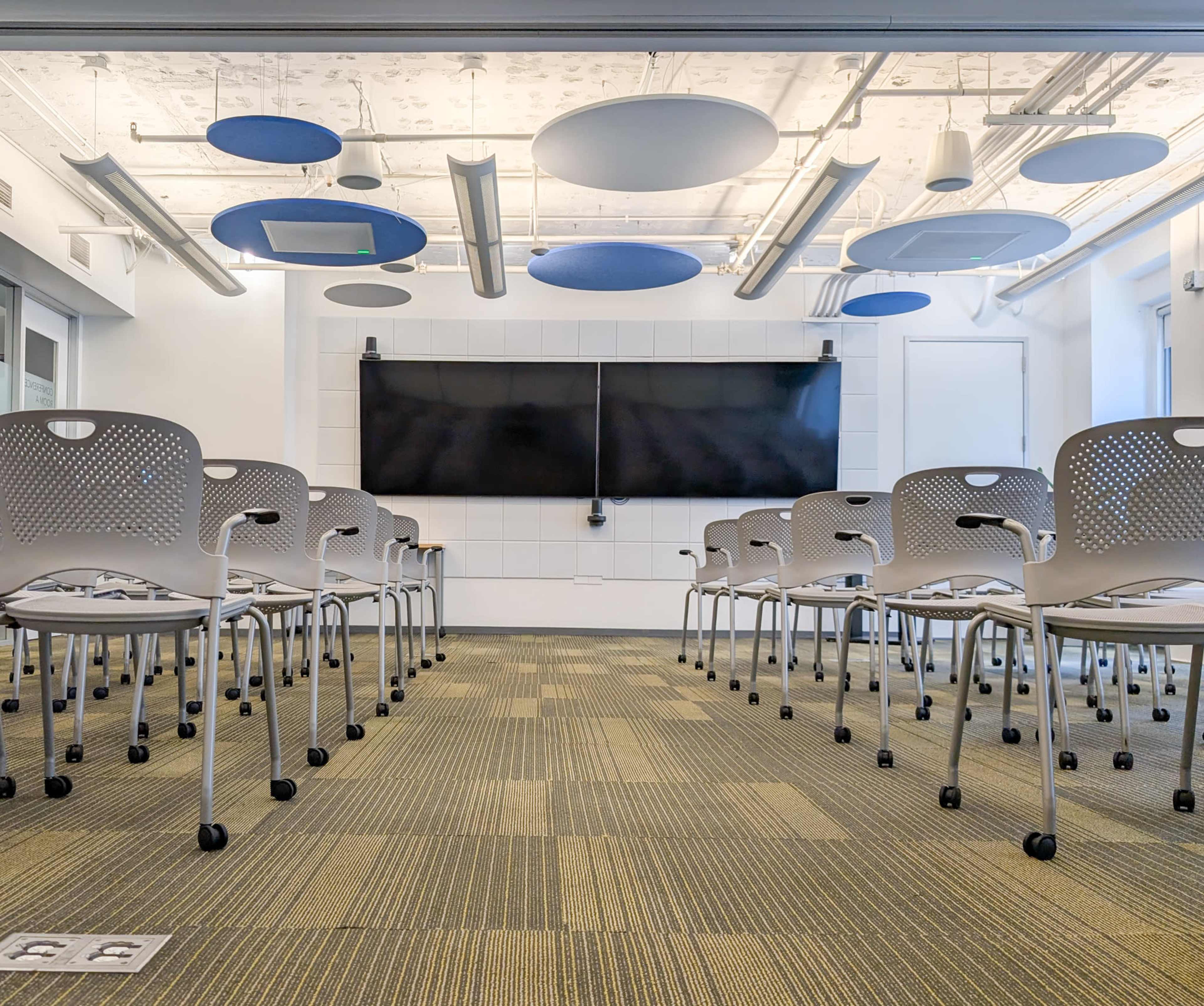The image shows a modern classroom setup with rows of white chairs facing a large black screen on a white wall, under a ceiling with acoustic panels.