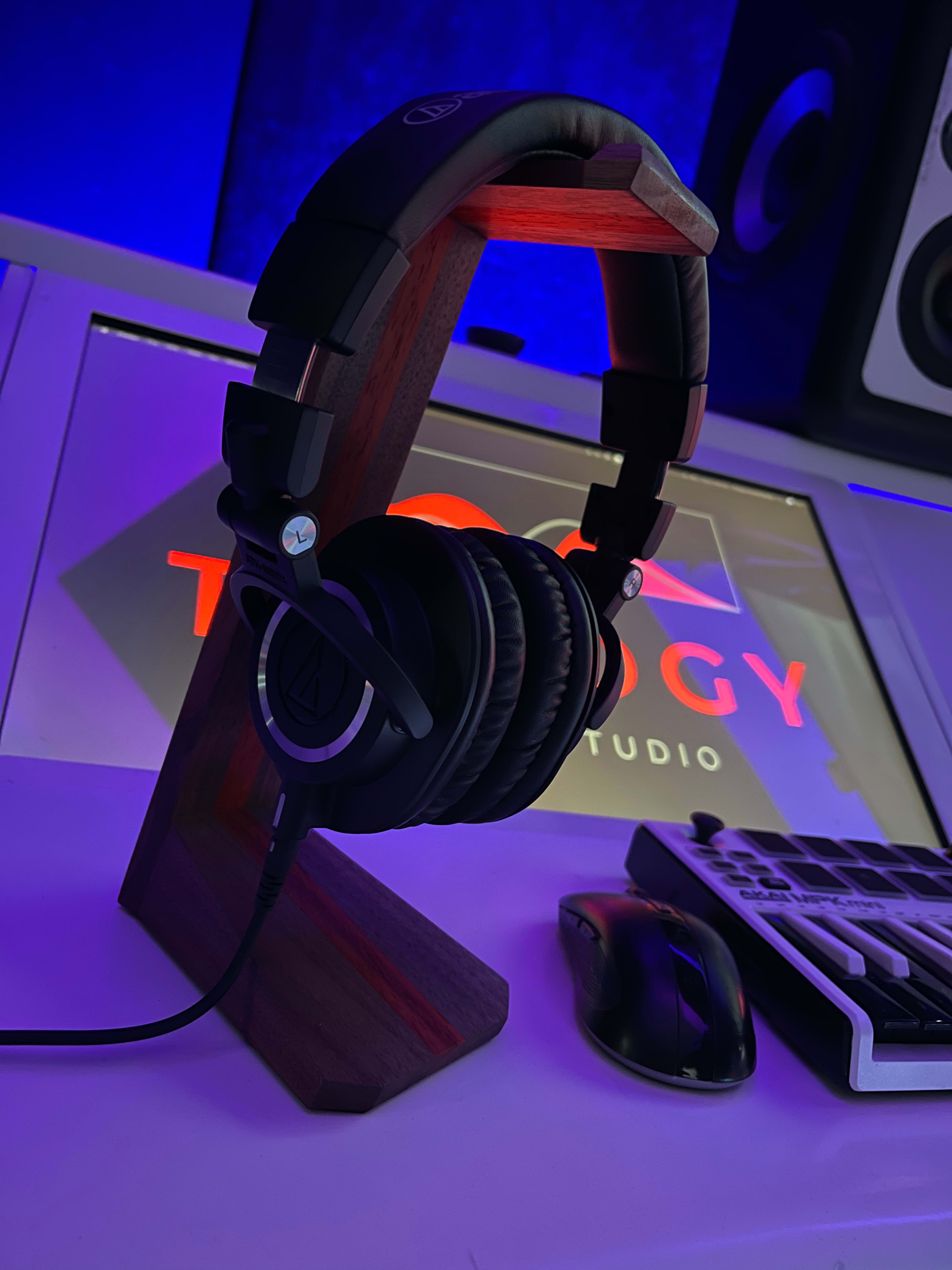 A pair of headphones is resting on a wooden stand in a studio workspace, with a backlit logo and a keyboard visible in the background.