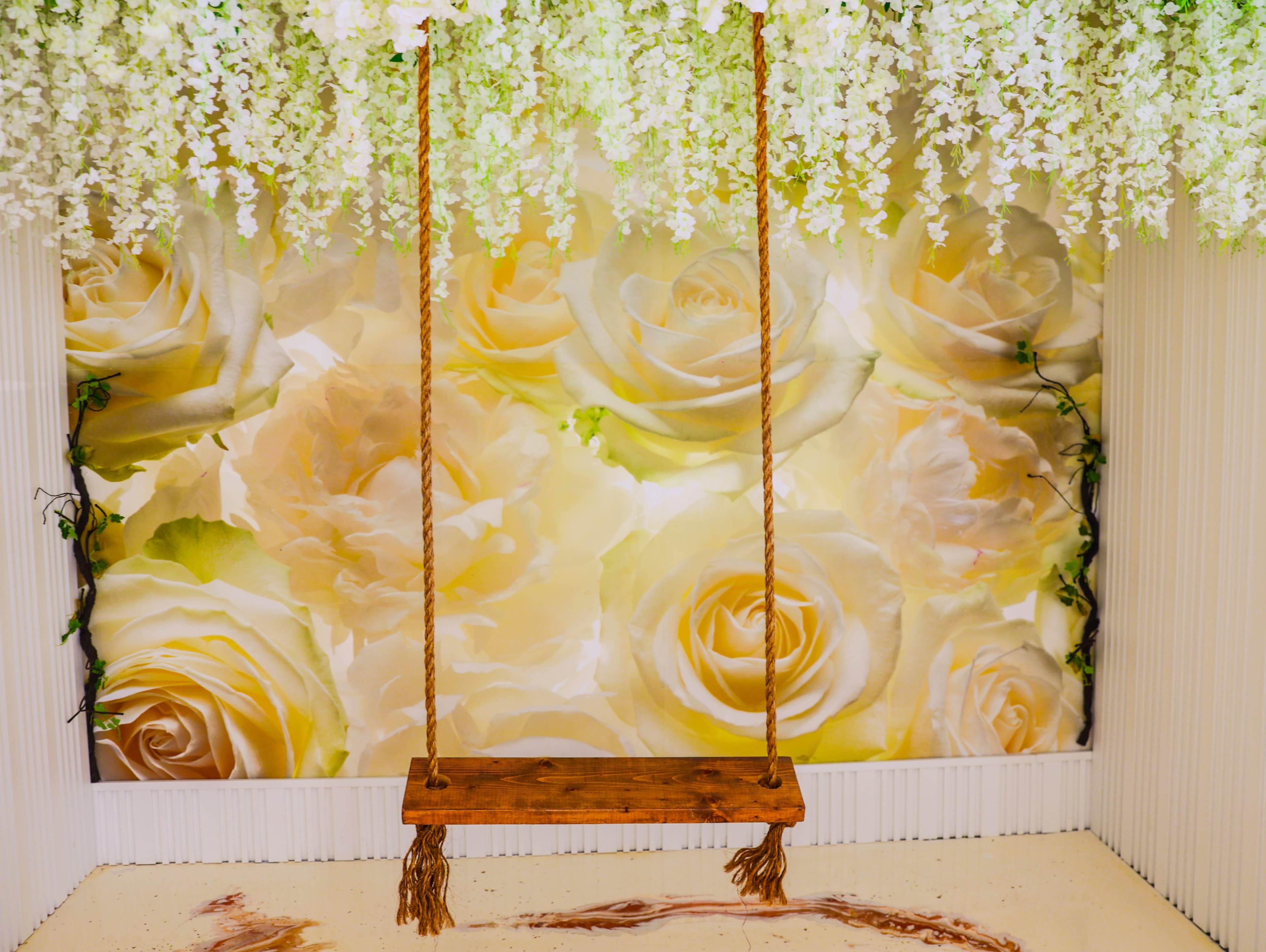 The image features a wooden swing hanging from a ceiling adorned with green foliage, set against a large, soft-focus backdrop of yellow roses.