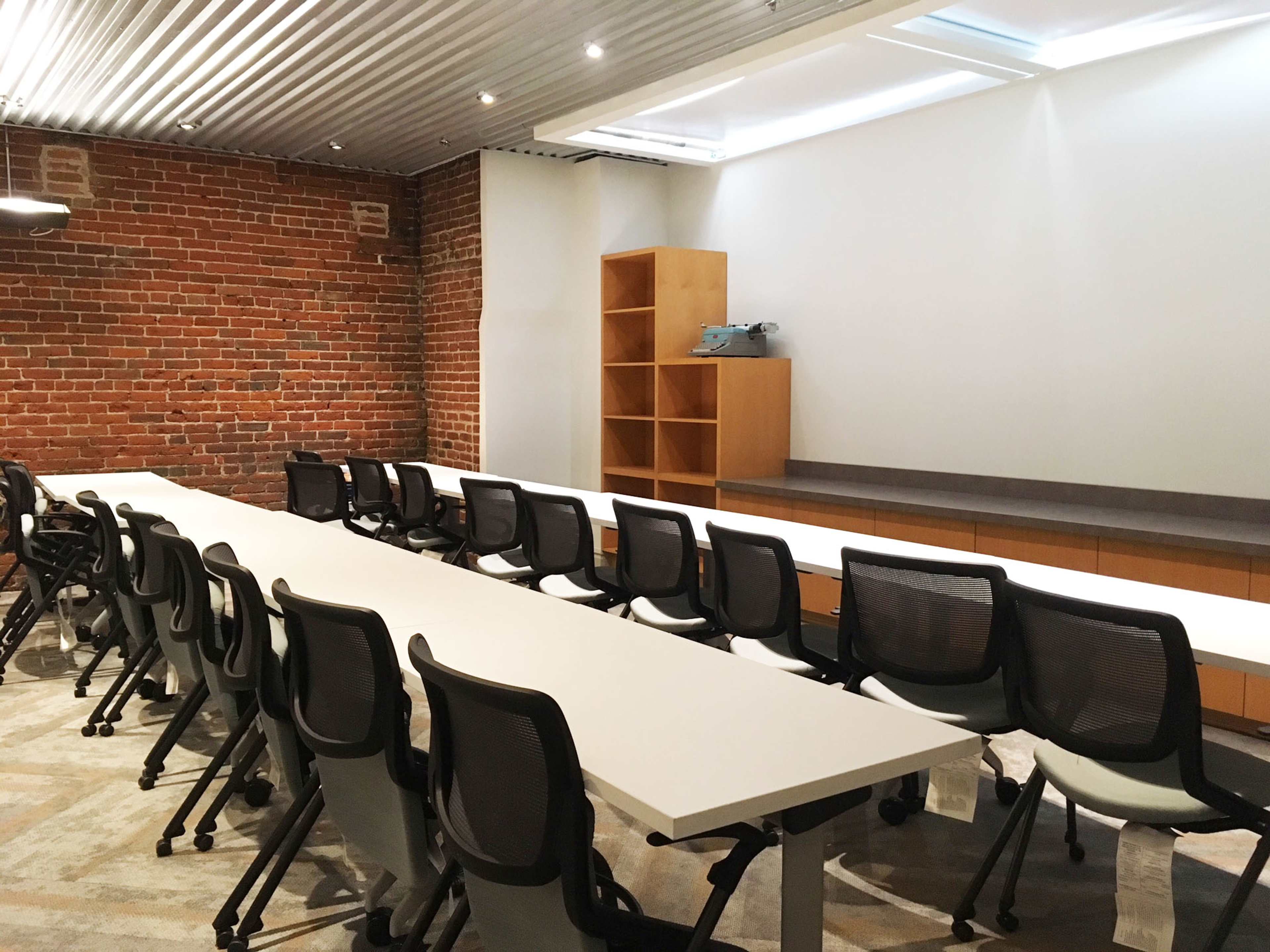 A modern meeting room with a long white table and several ergonomic chairs arranged in rows, along with a brick wall and wooden shelves in the background.