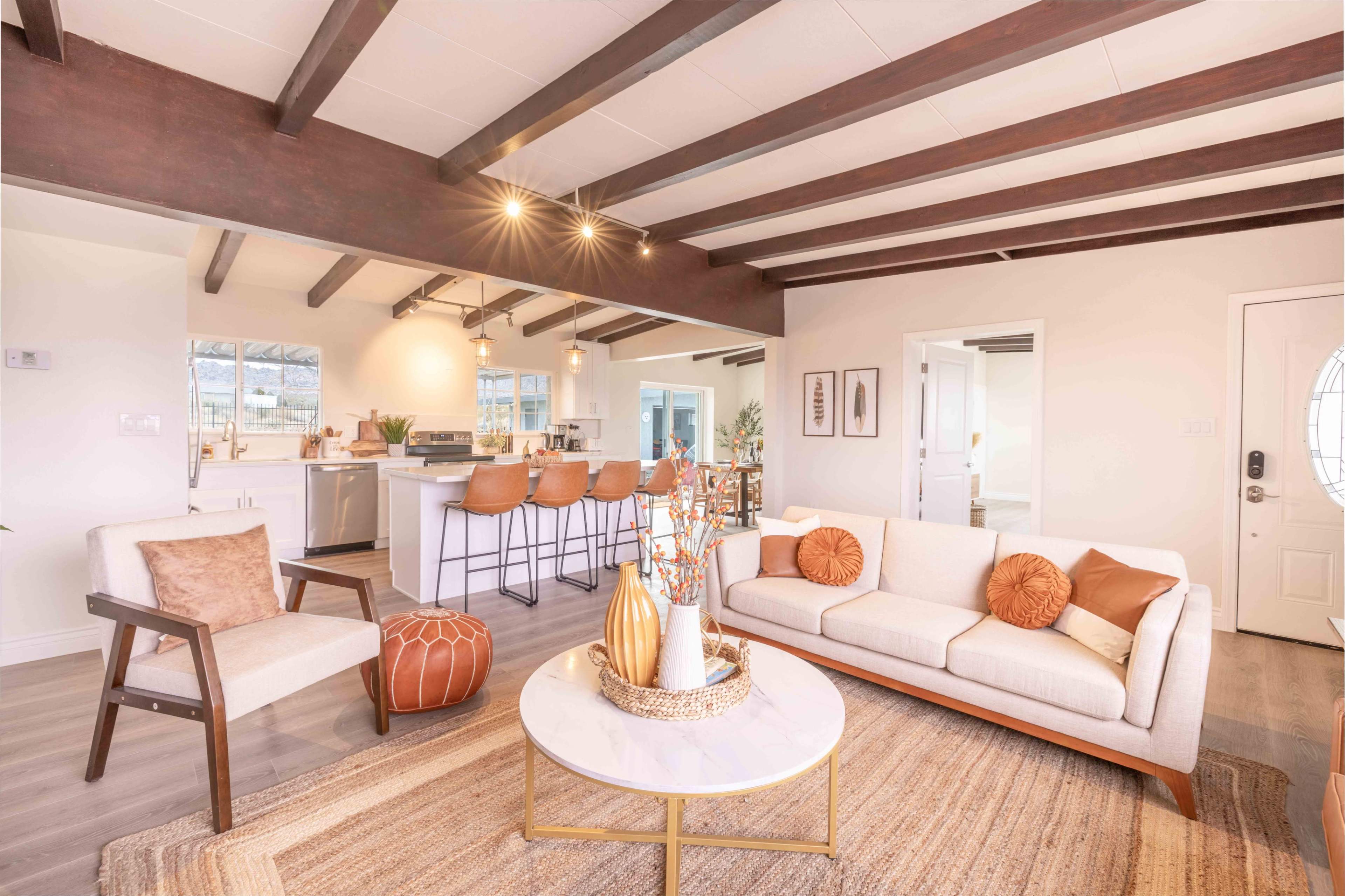 The image shows a modern living room with a beige sofa, a round coffee table, and a kitchen visible in the background with bar stools.