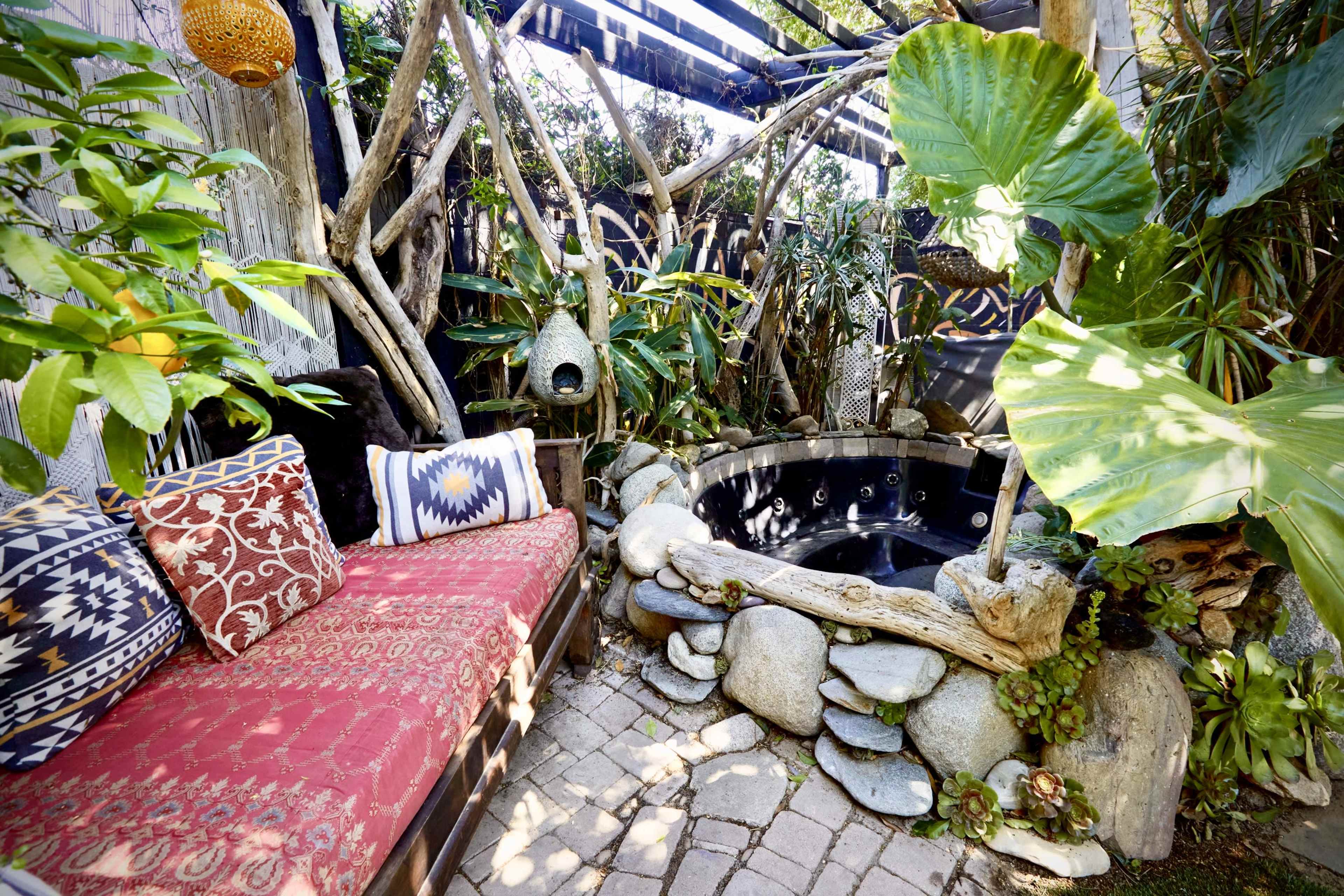 The image shows a cozy outdoor space featuring a circular stone hot tub surrounded by tropical plants and a rustic seating area with patterned cushions.