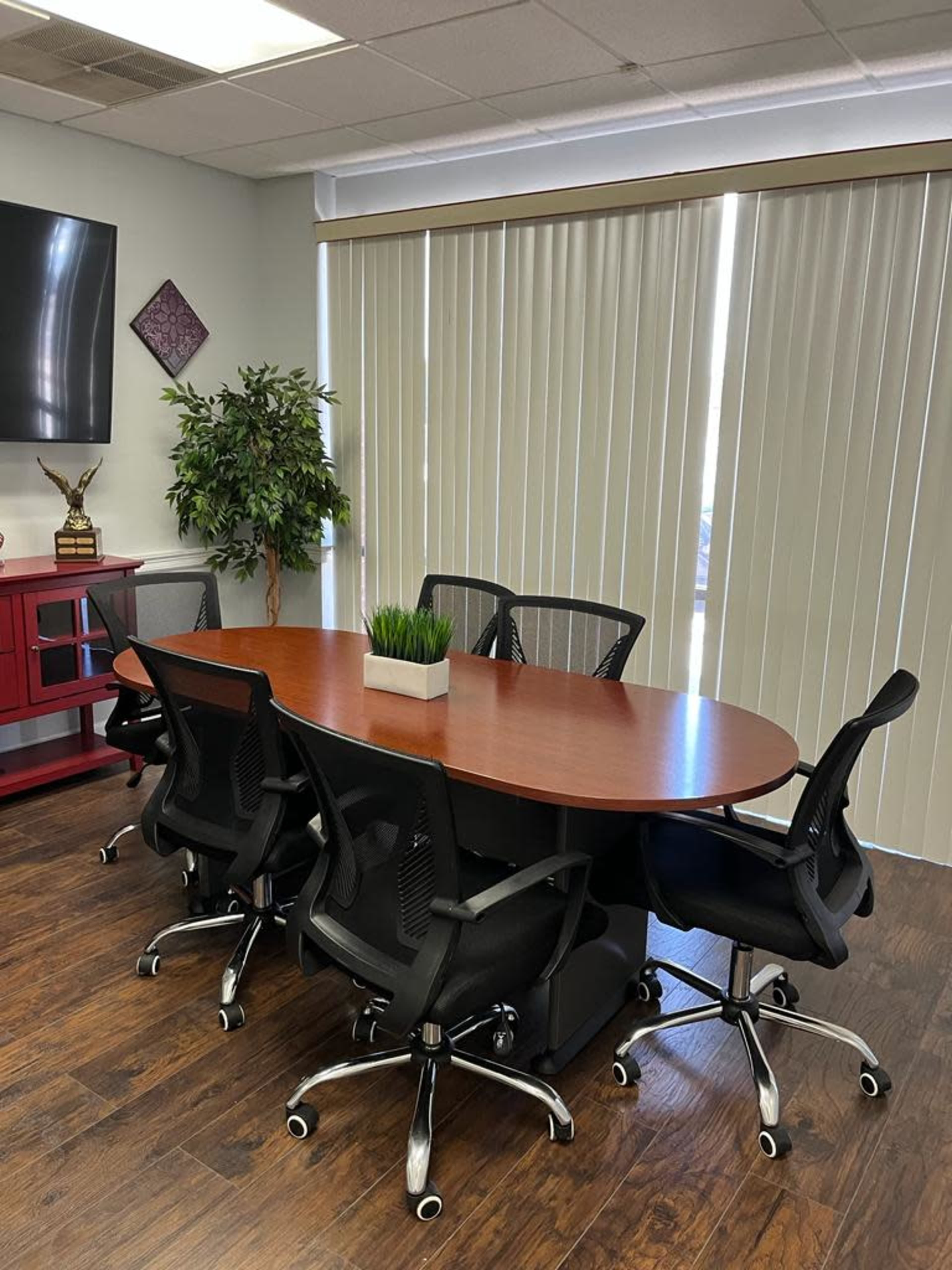 A conference room with a large oval wooden table surrounded by five black office chairs, a potted plant on the table, and large vertical blinds covering a window.
