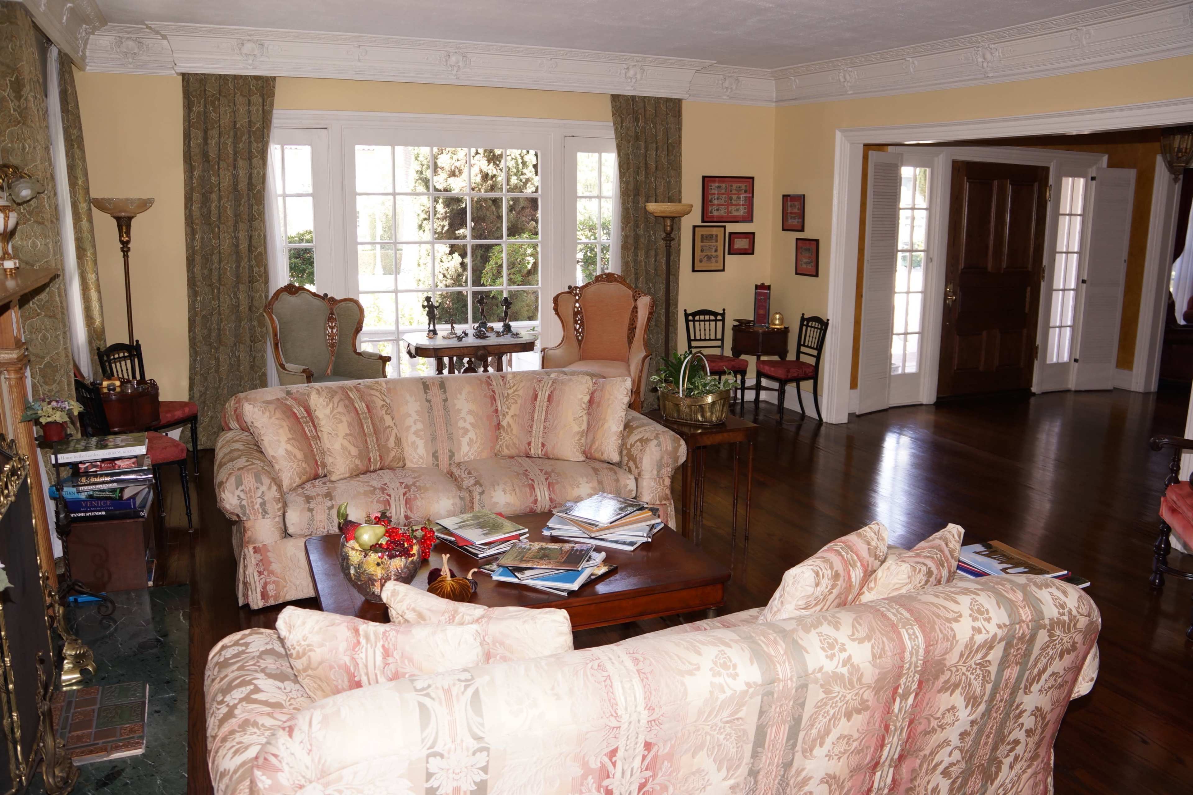 The image shows a spacious living room with two patterned couches, a wooden coffee table cluttered with magazines, and large windows letting in natural light.
