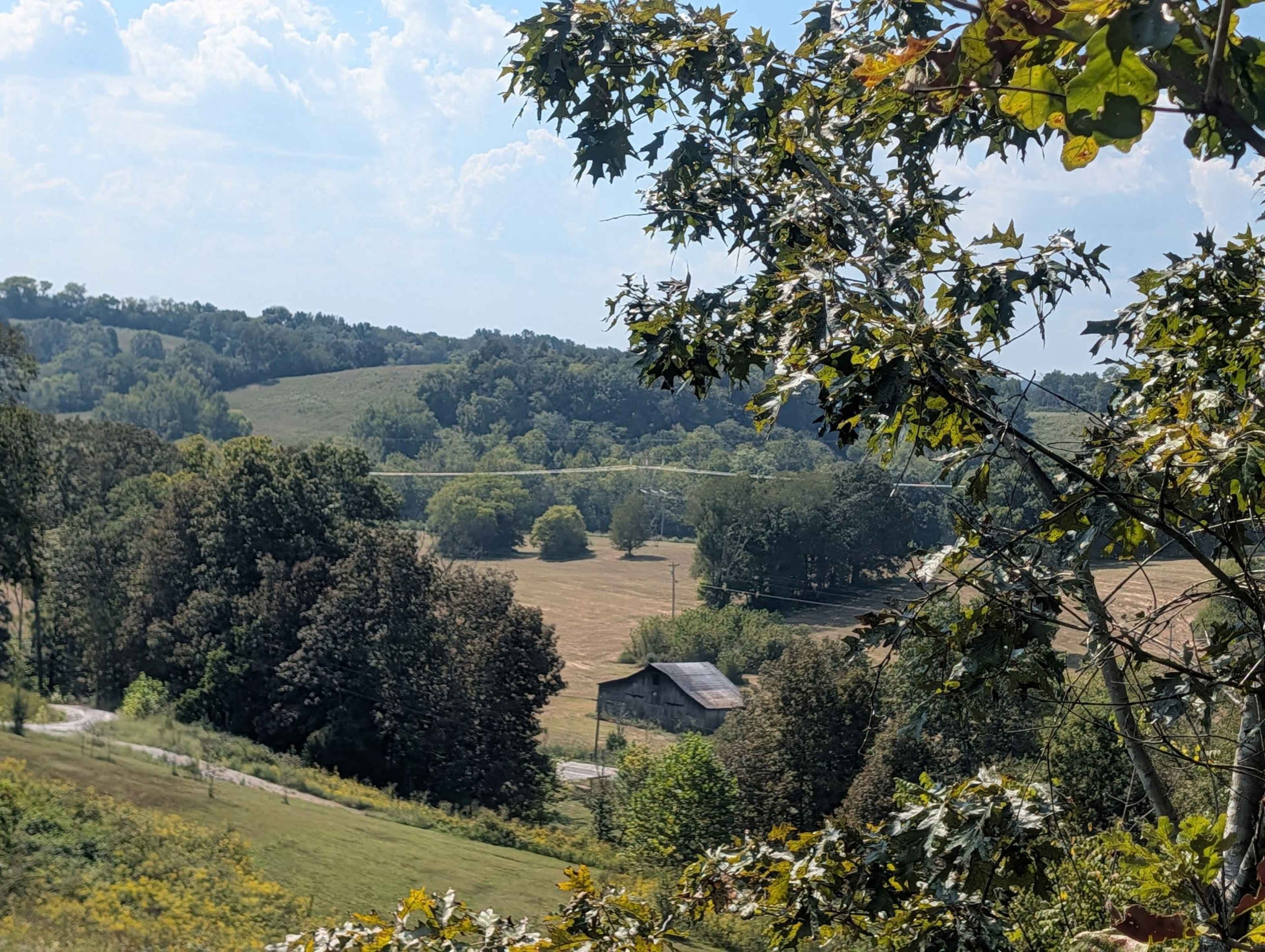 A rural landscape features rolling hills, a barn, and fields surrounded by trees under a partly cloudy sky.