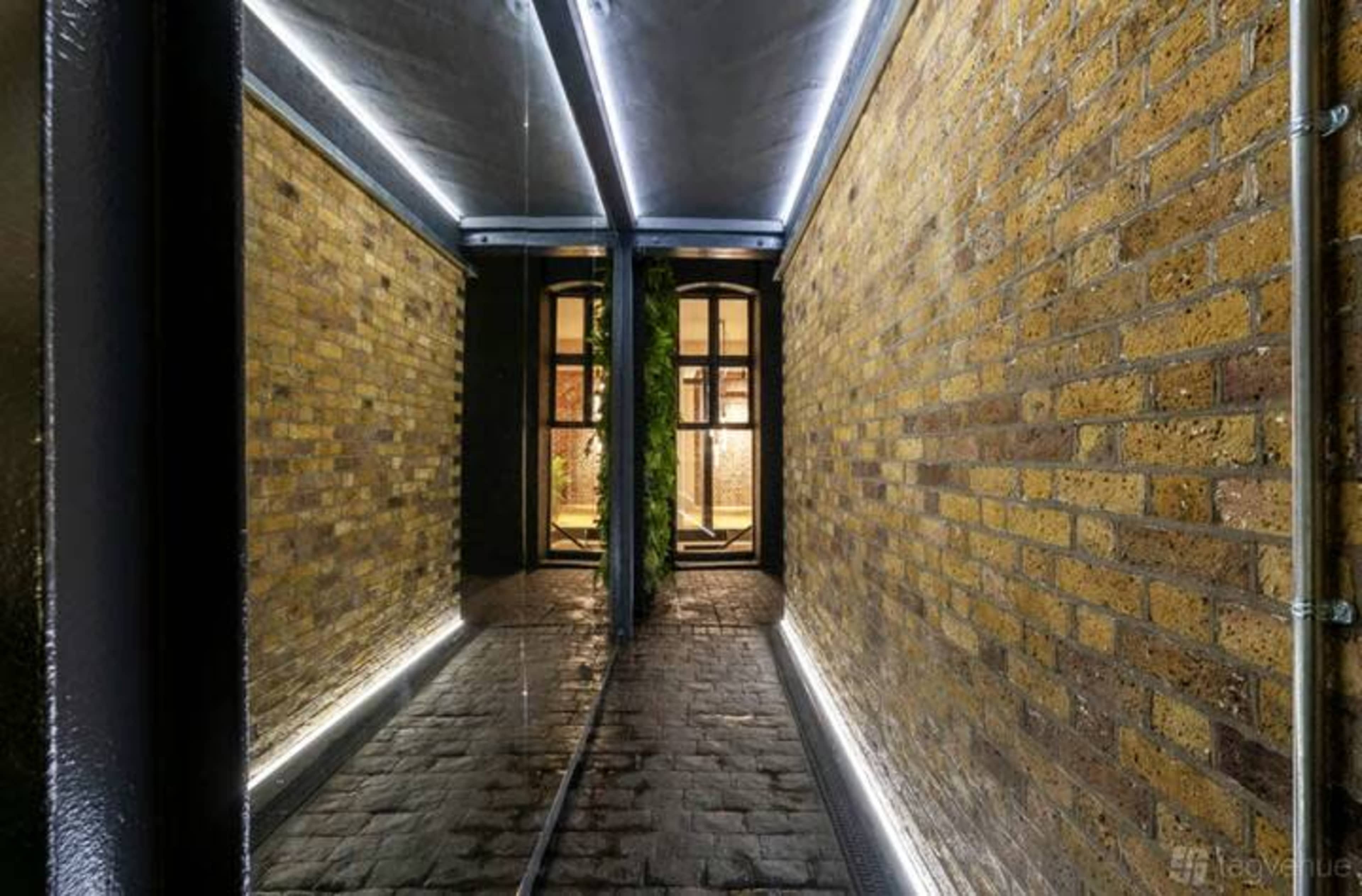 The image shows a narrow corridor with brick walls, illuminated by linear lighting, leading to a doorway that provides a glimpse of a well-lit room beyond.