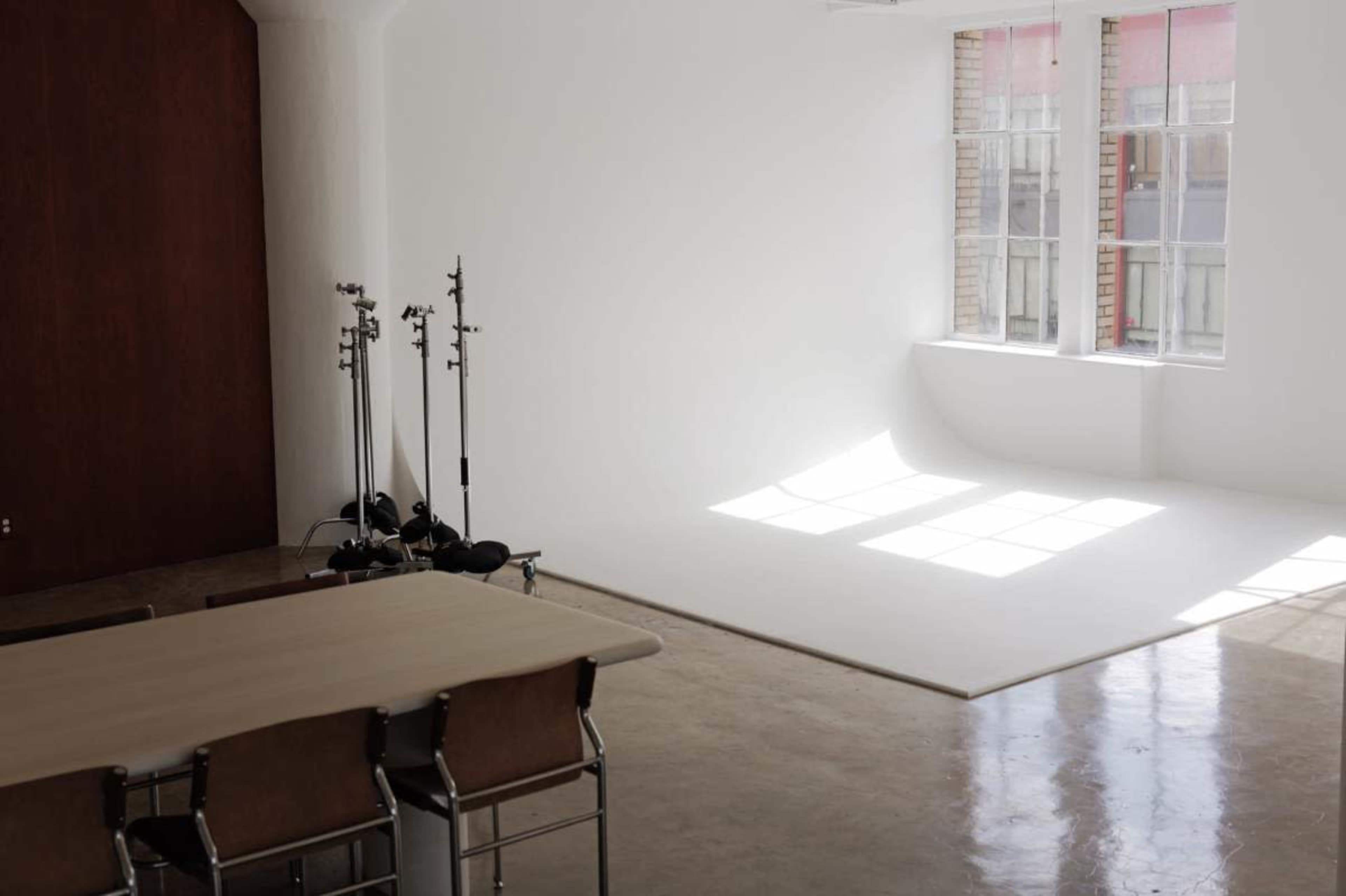 A bright, minimalist studio features a large white backdrop, two windows letting in natural light, and a wooden table with chairs.