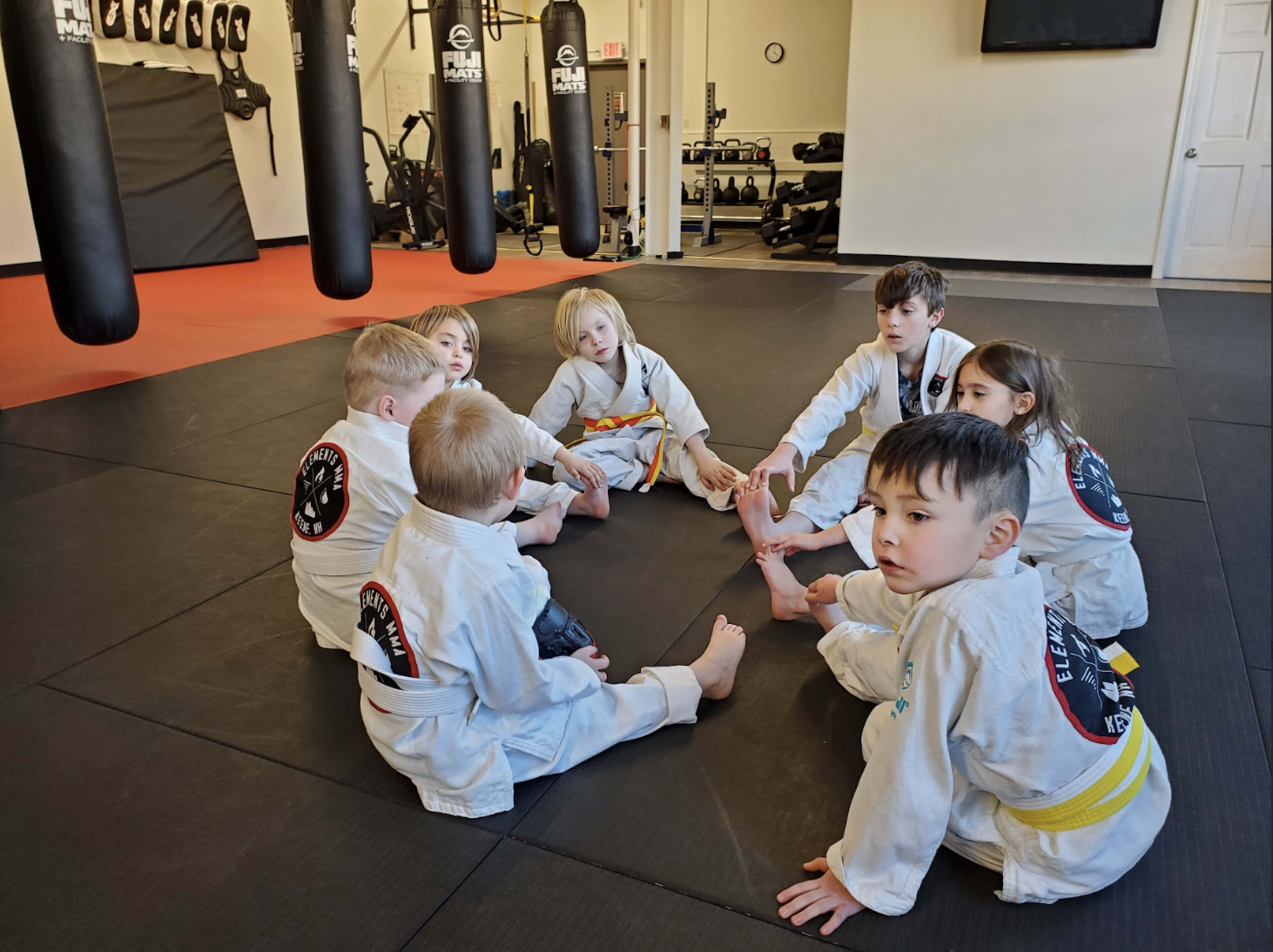 A group of children in martial arts uniforms sits in a circle on a black mat in a training facility.
