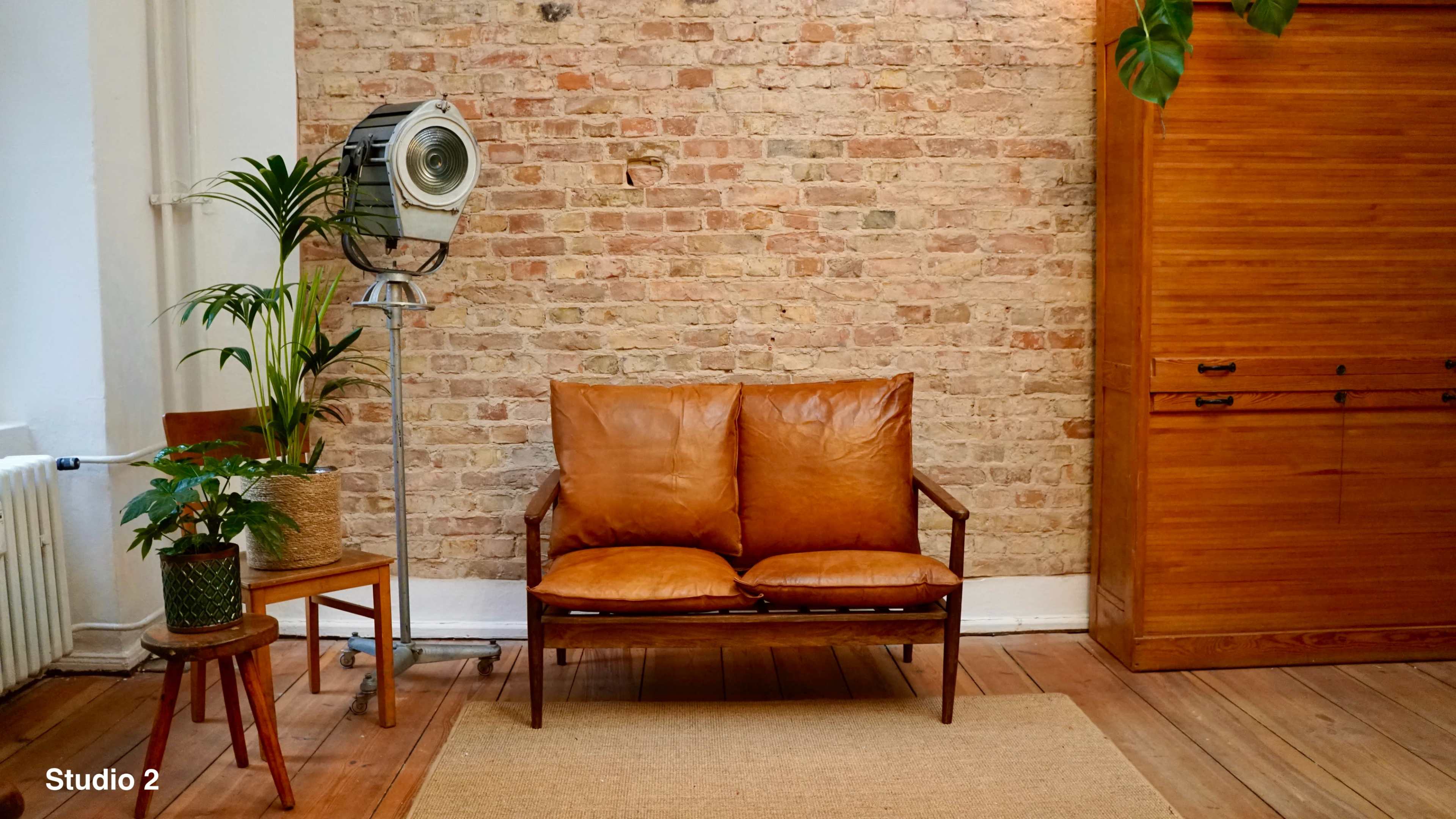 The image shows a cozy living space featuring a brown leather loveseat, a potted plant, a wooden chair, and a vintage floor lamp against a brick wall.
