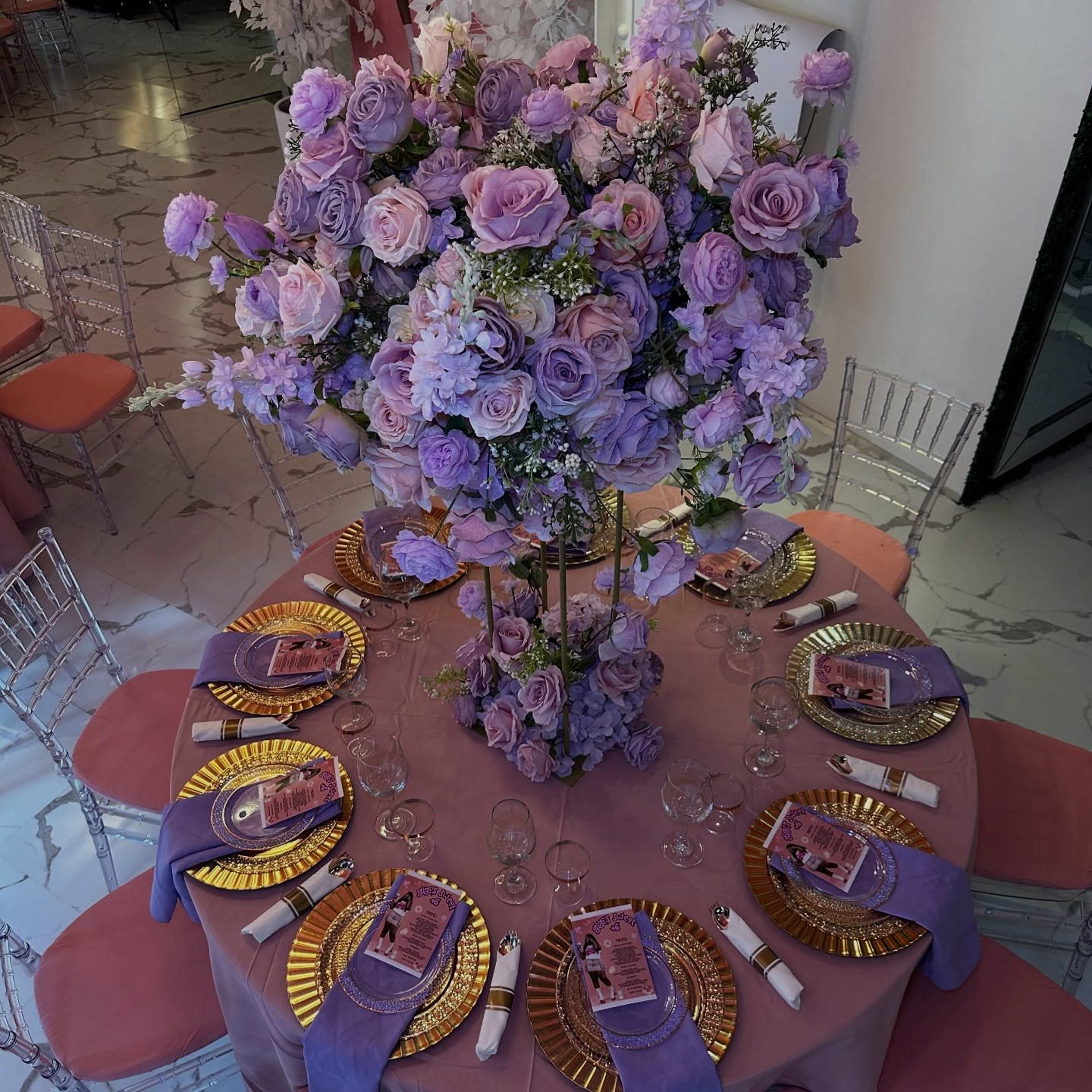A round table is elegantly set with gold-rimmed plates, lavender napkins, and topped with a tall floral arrangement of purple roses and other flowers.