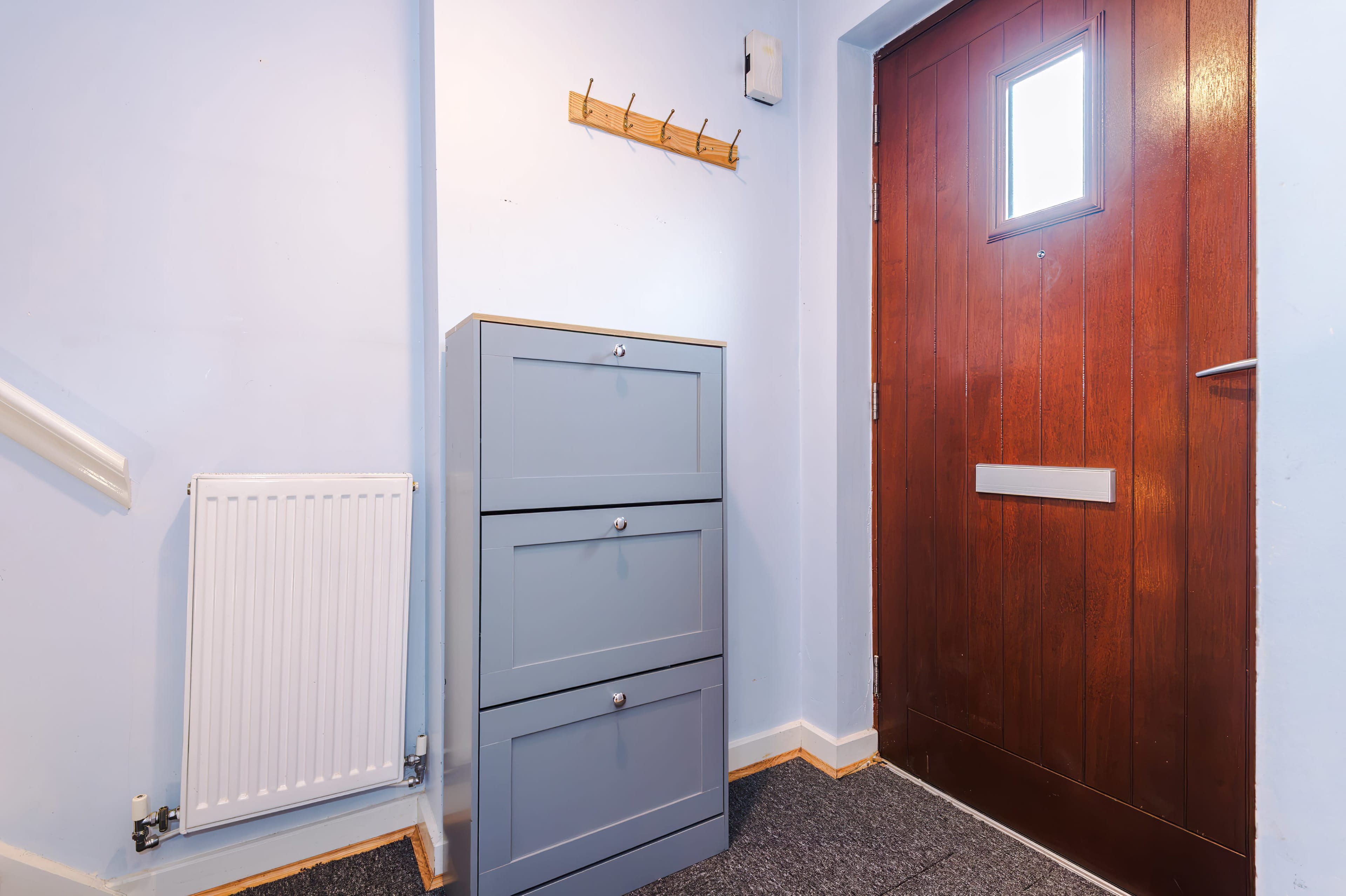 The image shows an entryway with a wooden front door, a small gray cabinet with three drawers, a radiator, and a coat hook on a light blue wall.