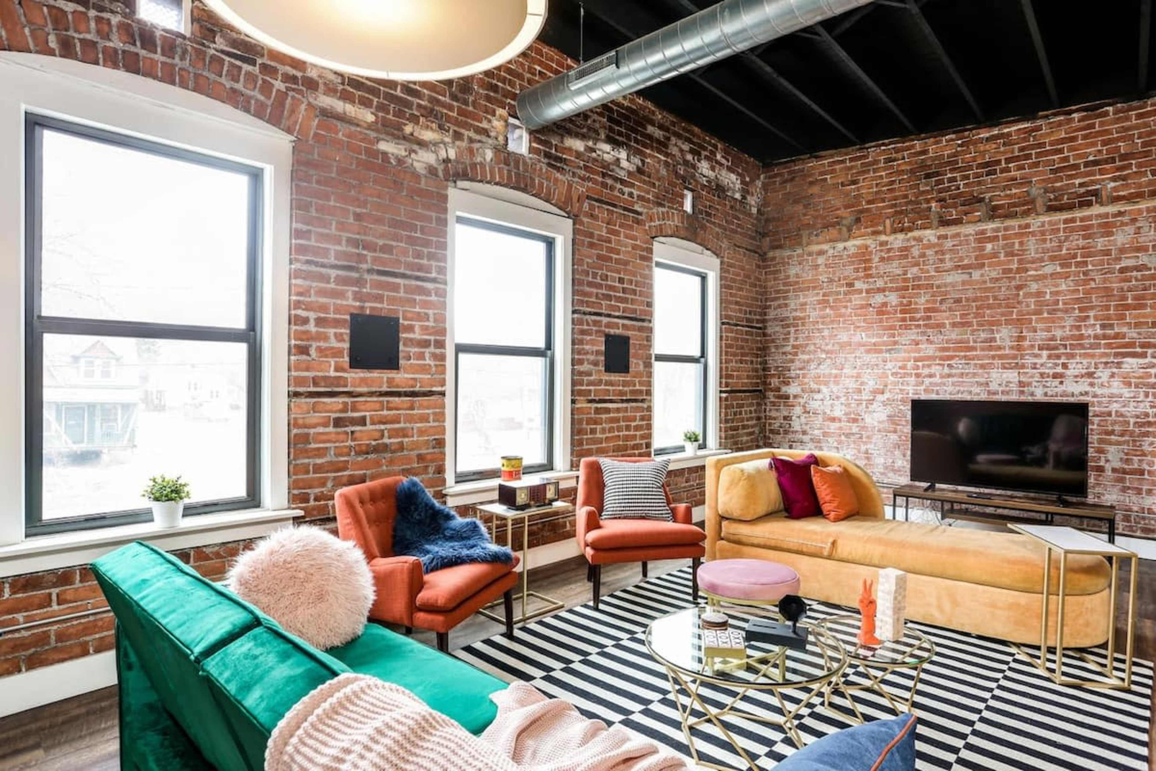 A modern living room with exposed brick walls, large windows, and colorful furniture arranged around a striped rug.