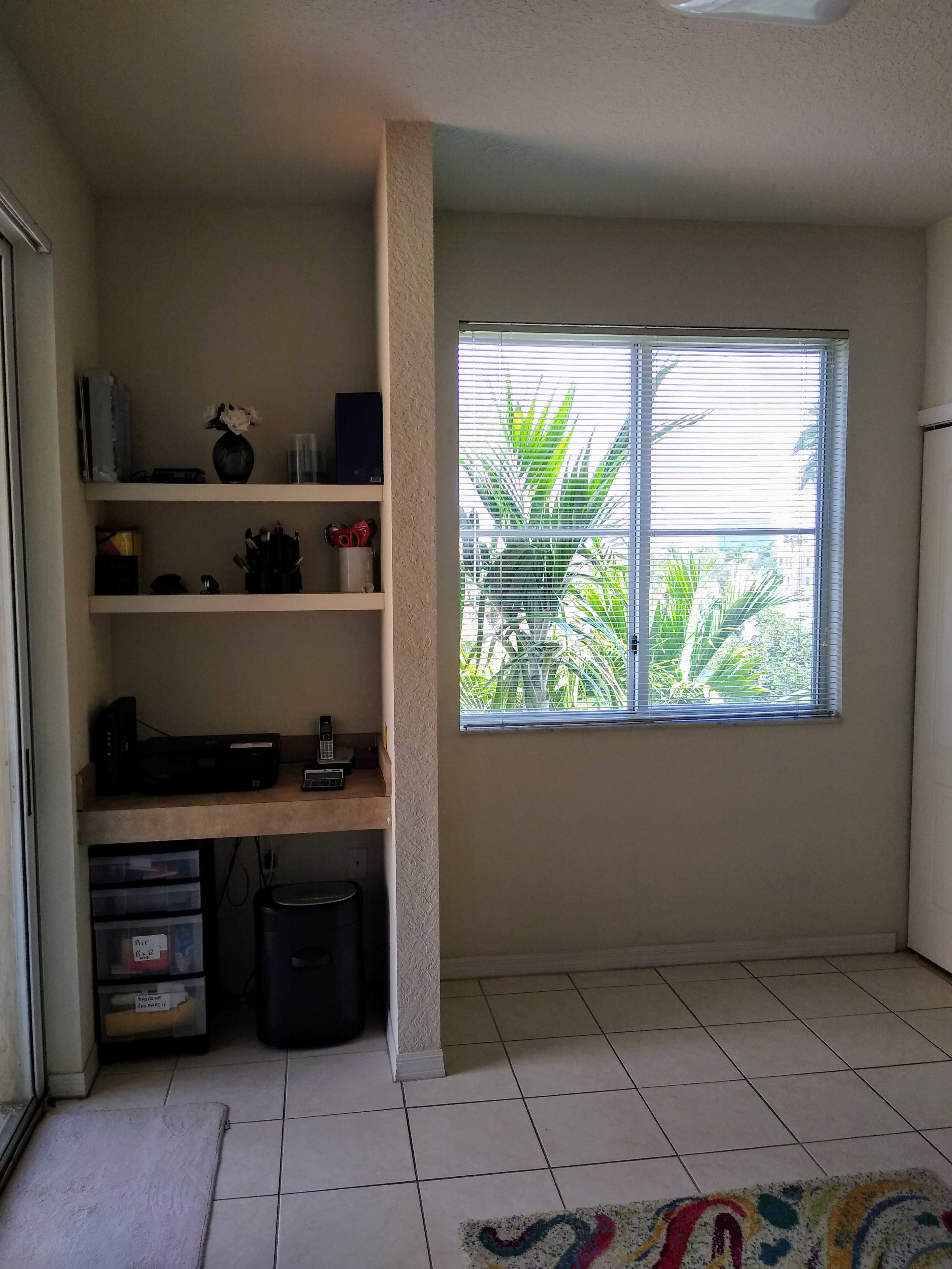 A small, well-lit room features a desk with office supplies and a window overlooking a palm tree landscape.