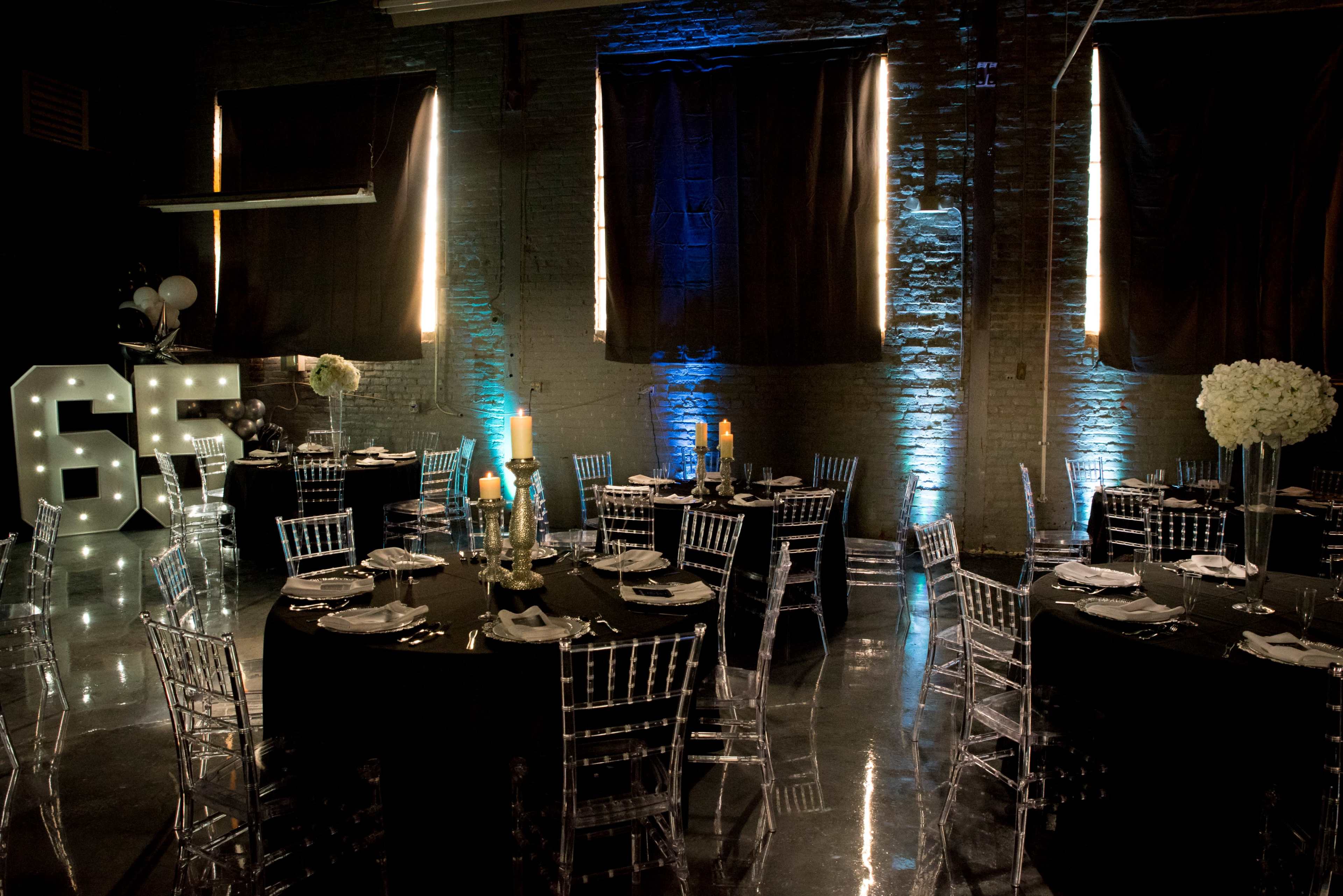 The scene features a banquet hall set for a celebration, with round tables covered in black cloth, clear Chiavari chairs, and illuminated number displays reading "65."