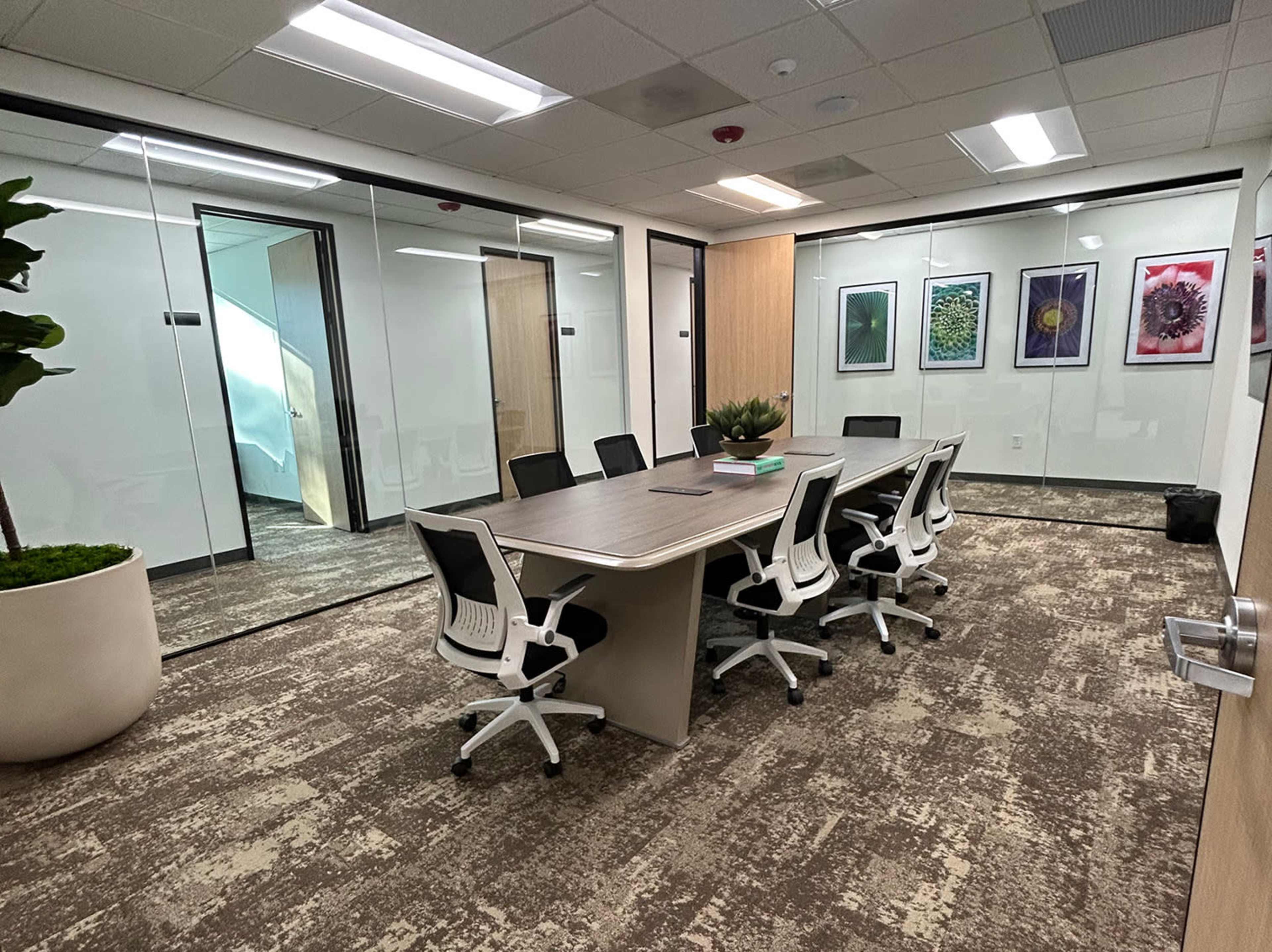 A modern conference room features a large table surrounded by white office chairs, with glass walls and framed artwork on display.