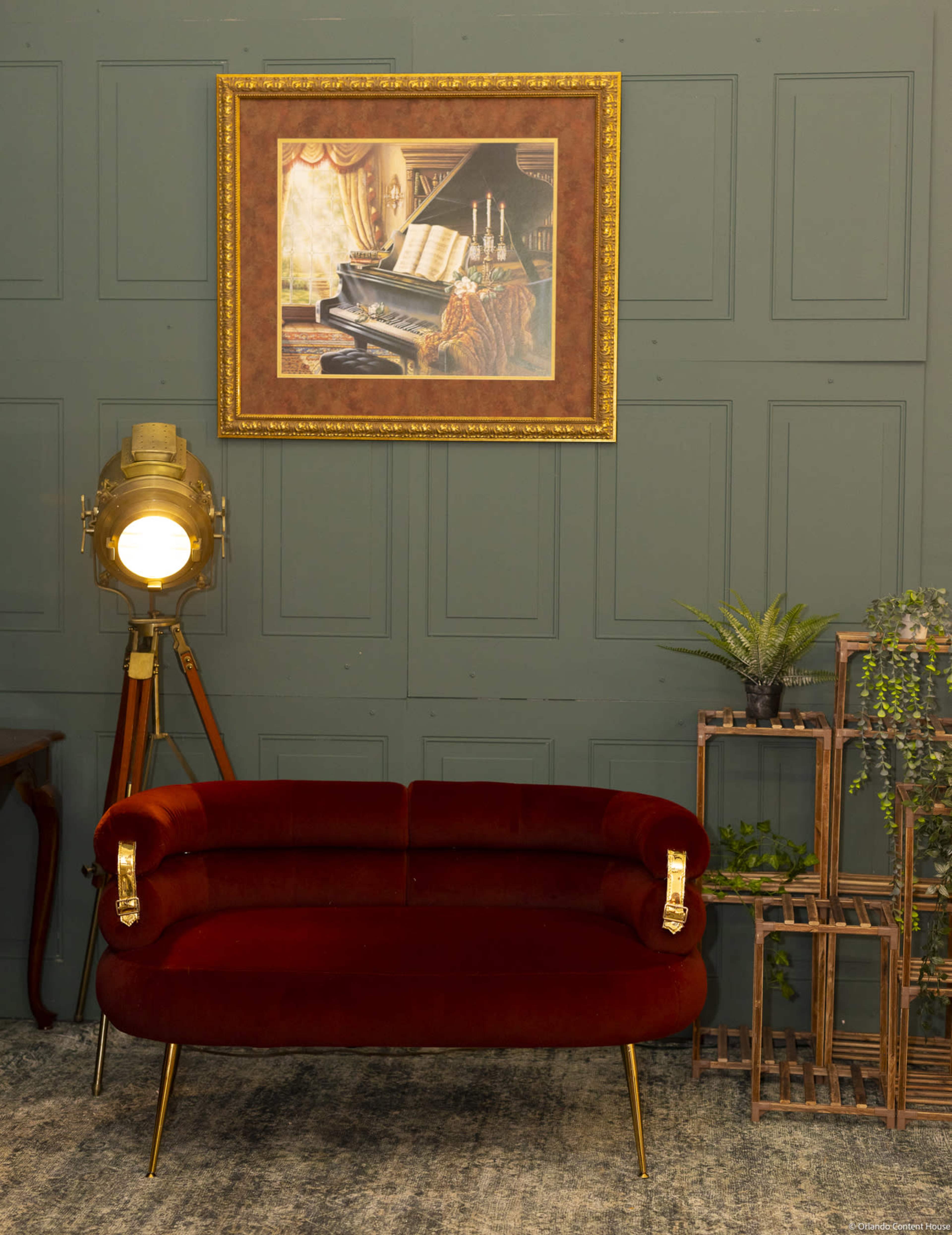 The image features a red velvet sofa positioned against a green paneled wall, with a framed painting of a piano above it and a vintage floor lamp nearby.