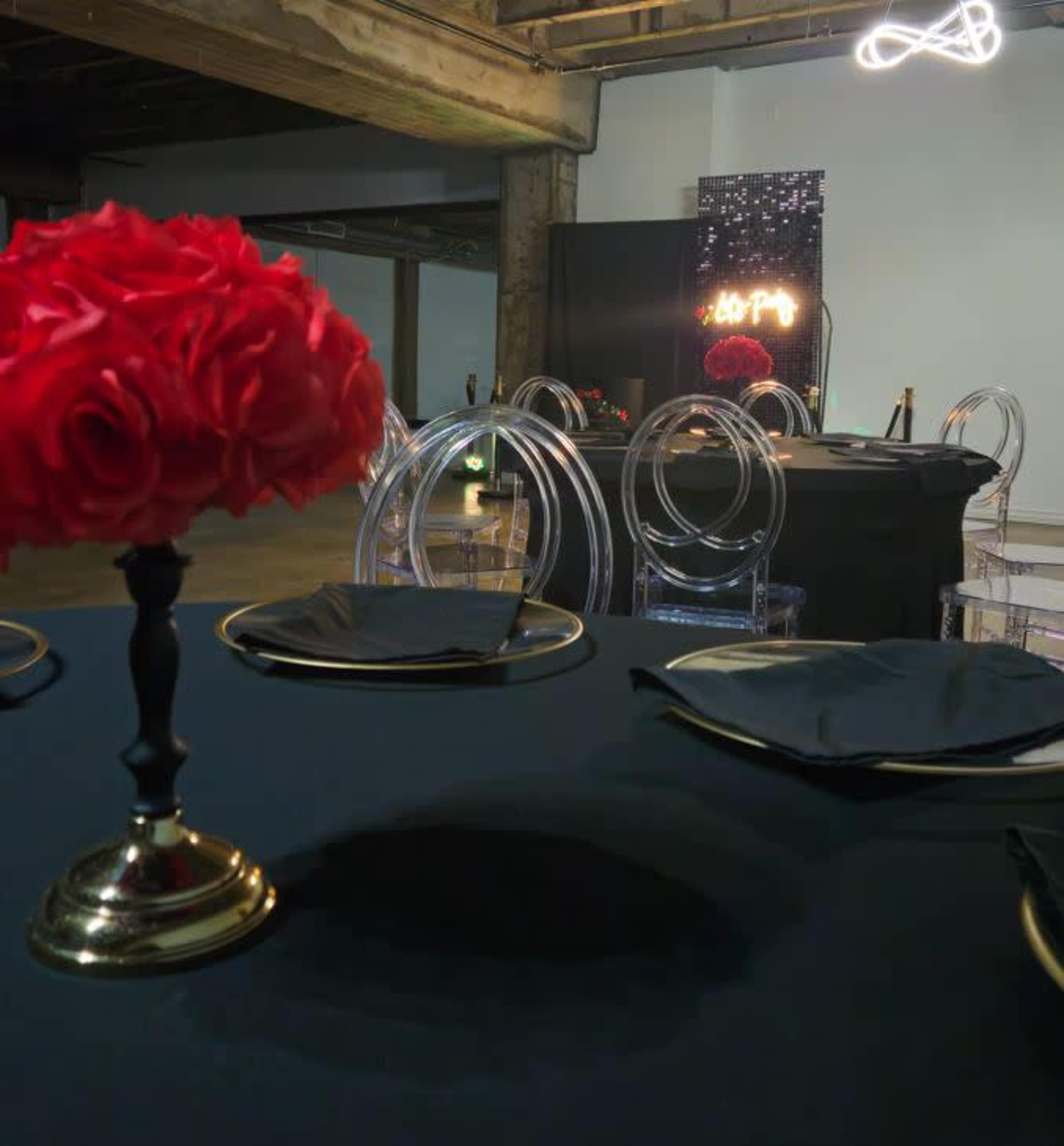 A decorated dining space features a black tablecloth, clear acrylic chairs, a centerpiece of red roses, and a neon sign in the background.