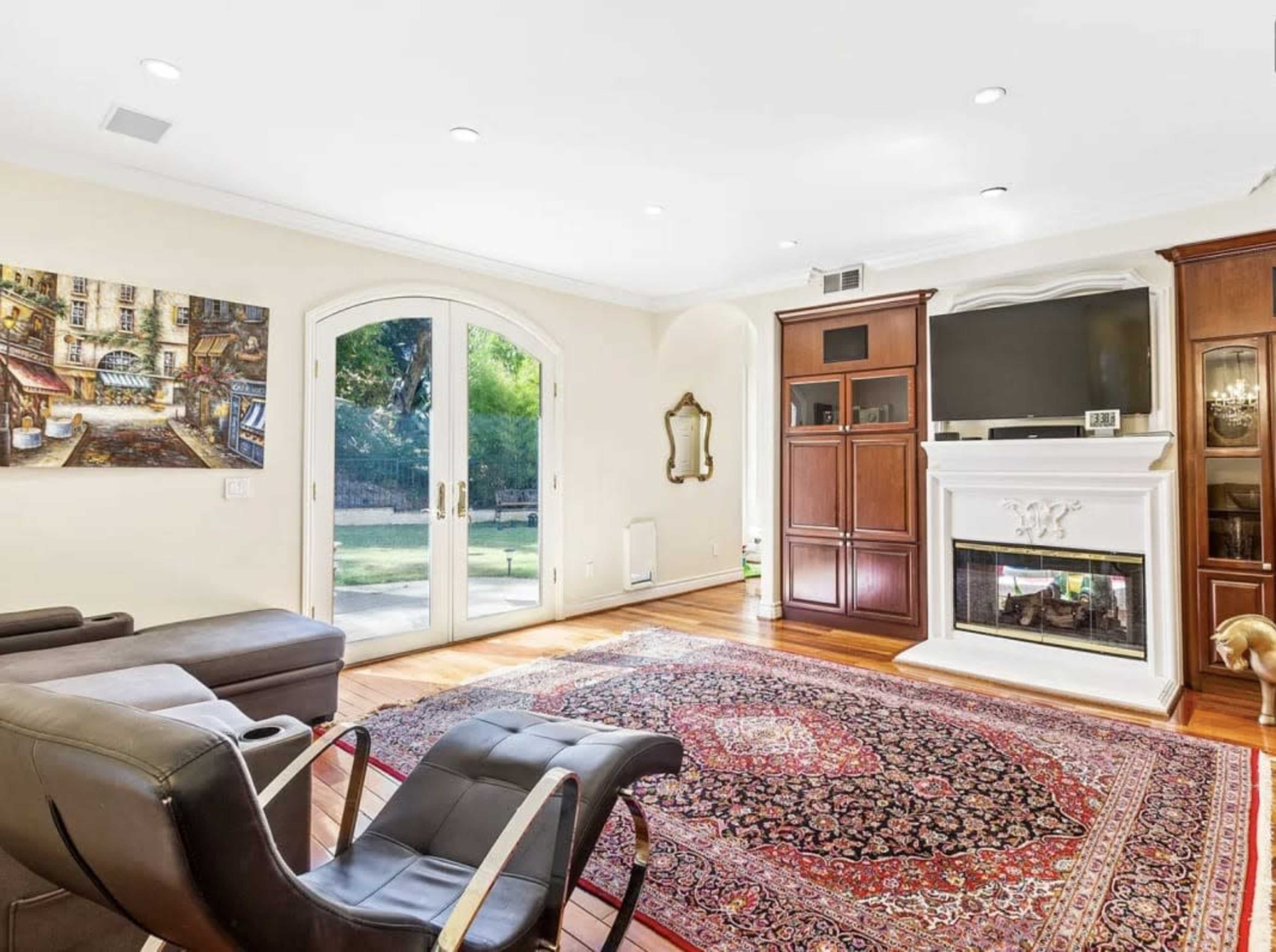 A cozy living room features a large area rug, a flat-screen TV mounted above a fireplace, and glass doors leading to a green outdoor space.