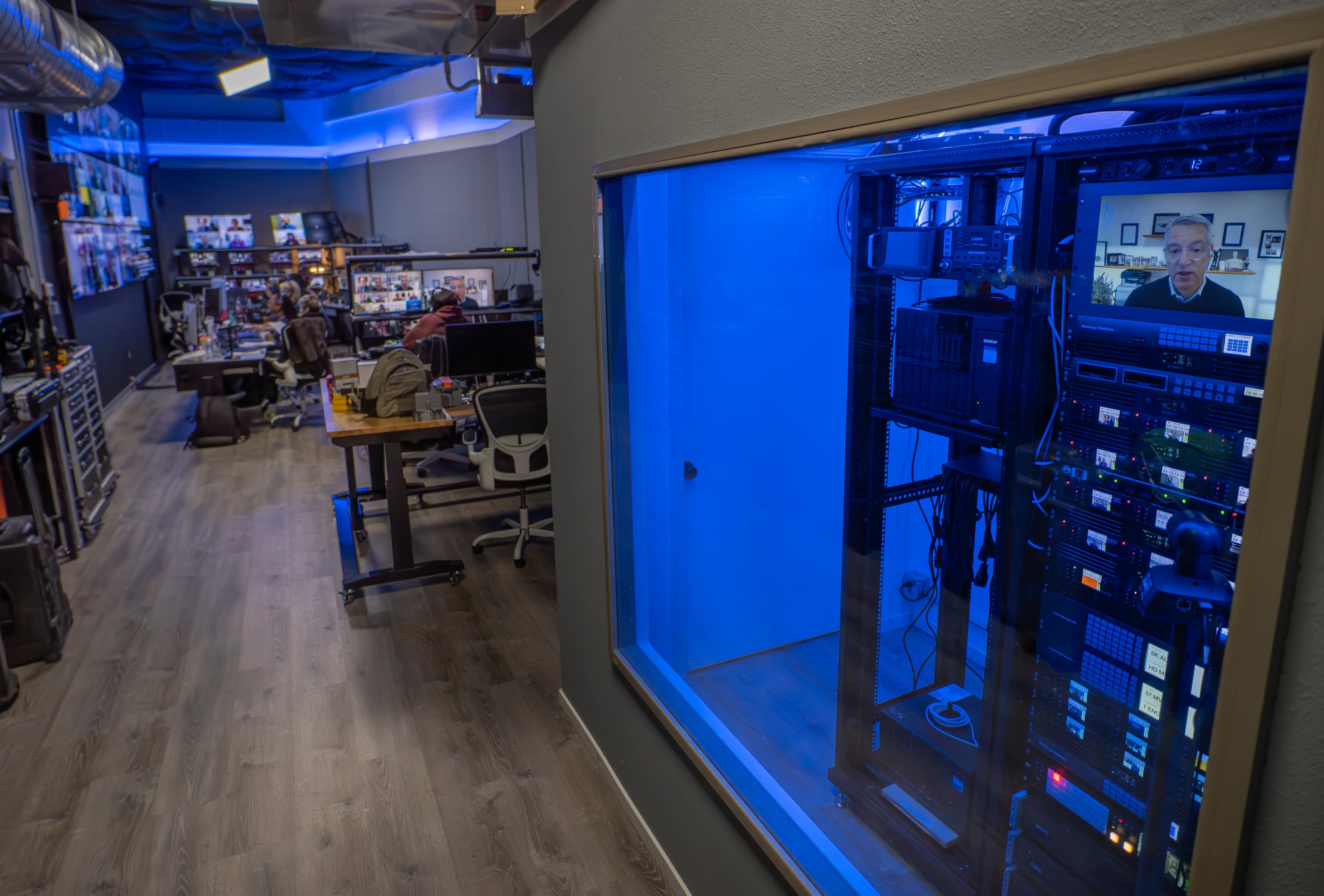 The image shows a modern office space with a server room featuring blue lighting on one side and workstations on the other, populated by multiple people.