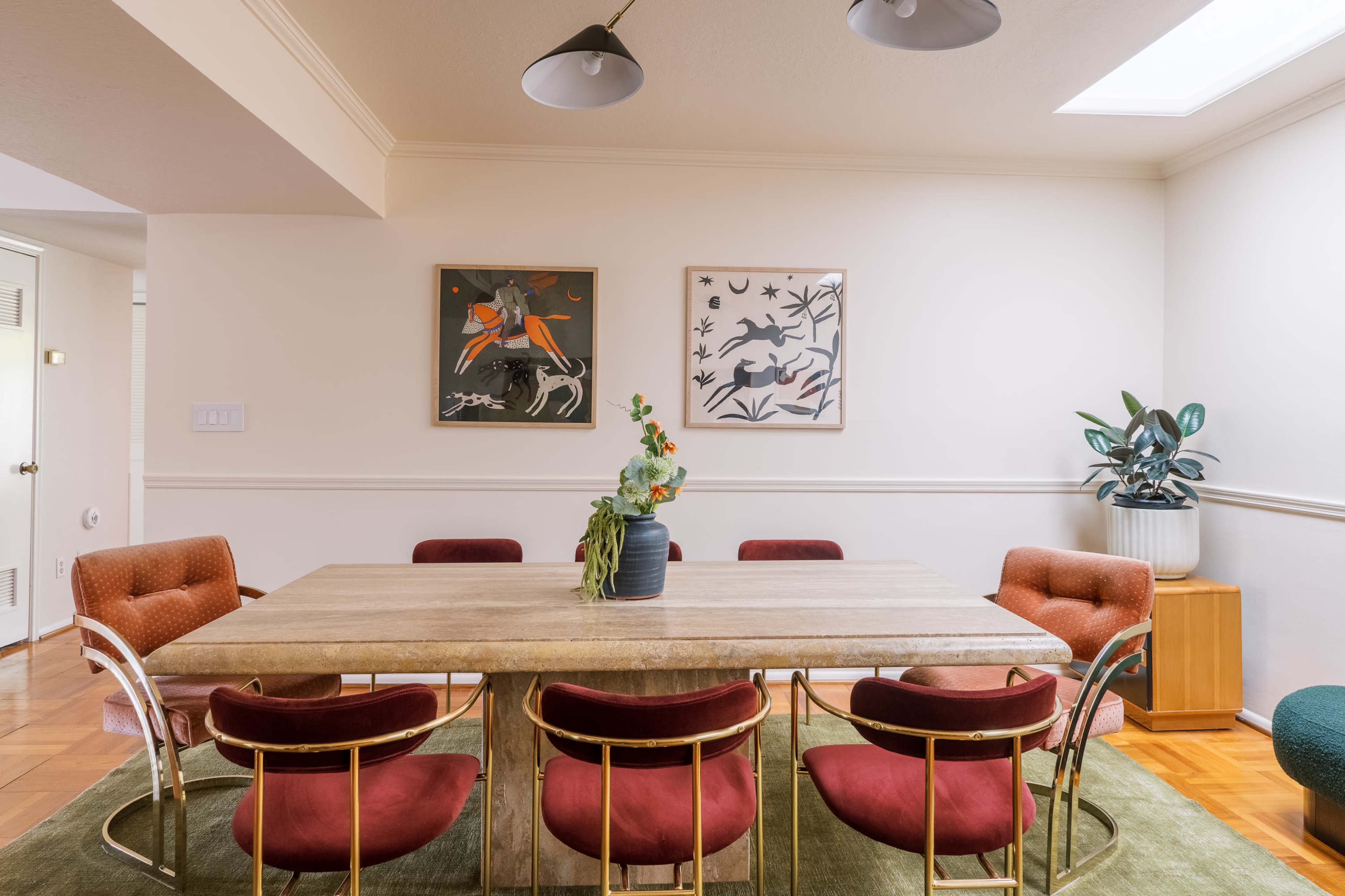 A dining room features a large rectangular table surrounded by eight chairs, with two framed artworks on the wall and a potted plant in the corner.