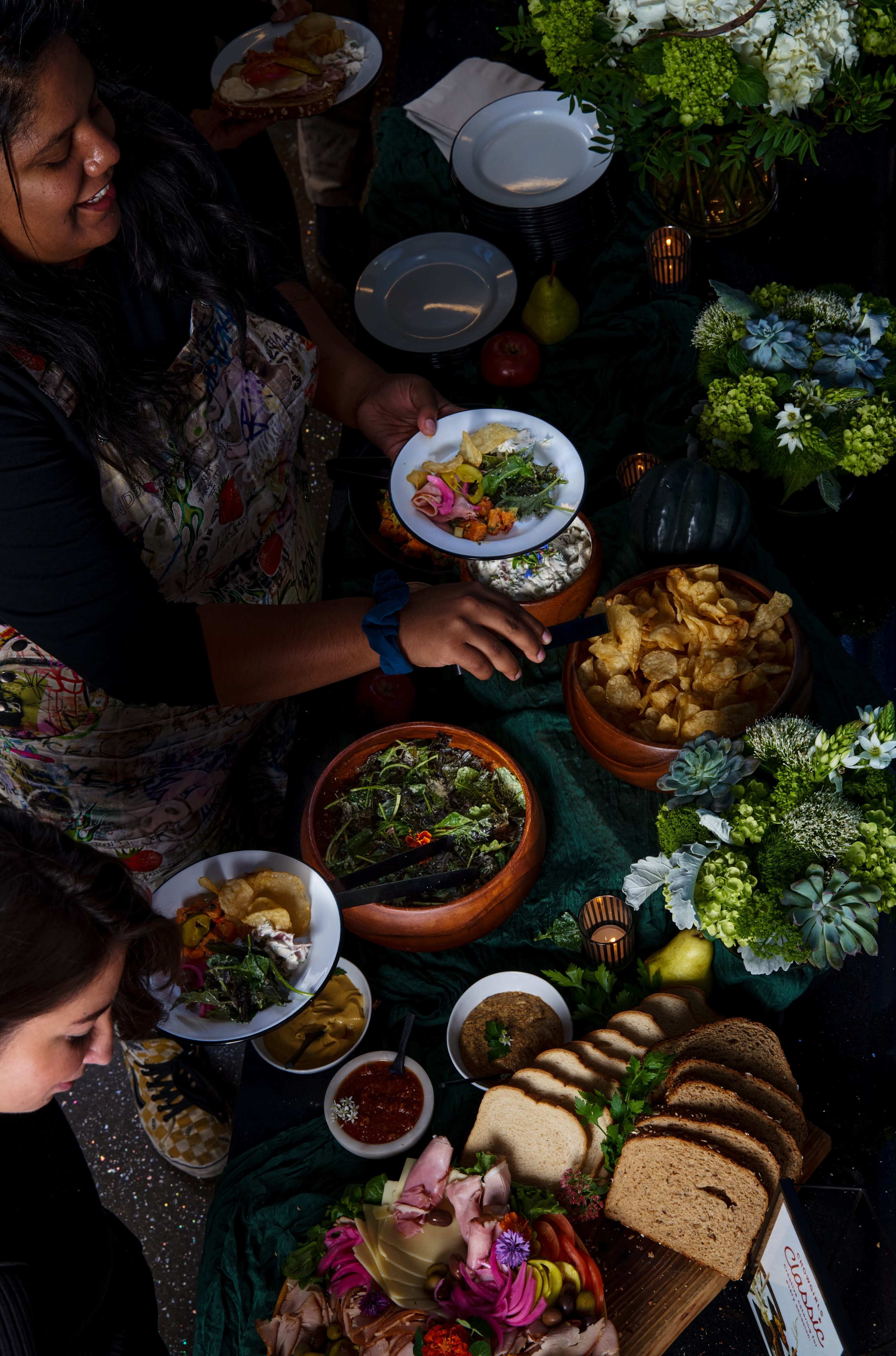 A woman serves food from a table featuring various dishes, including salads, bread, and snacks, surrounded by decorative elements like flowers and fruits.